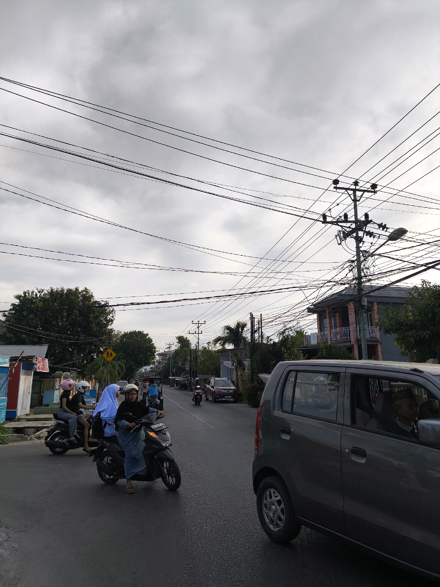 The image depicts a street scene in Gorontalo, Indonesia, under an overcast sky filled with visible power lines. The road is paved and appears wet, suggesting recent rain. On the left side of the road, there are small buildings and a yellow traffic sign depicting children crossing. Several motorcycles are present, with individuals on them, some wearing helmets. One motorcycle in the foreground carries three people, including a woman in a blue hijab. A dark grey car is visible in the foreground on the right, also on the road. Trees and some buildings with balconies are situated along the sides of the street. The overall impression is of a typical urban street environment in Indonesia with active traffic.