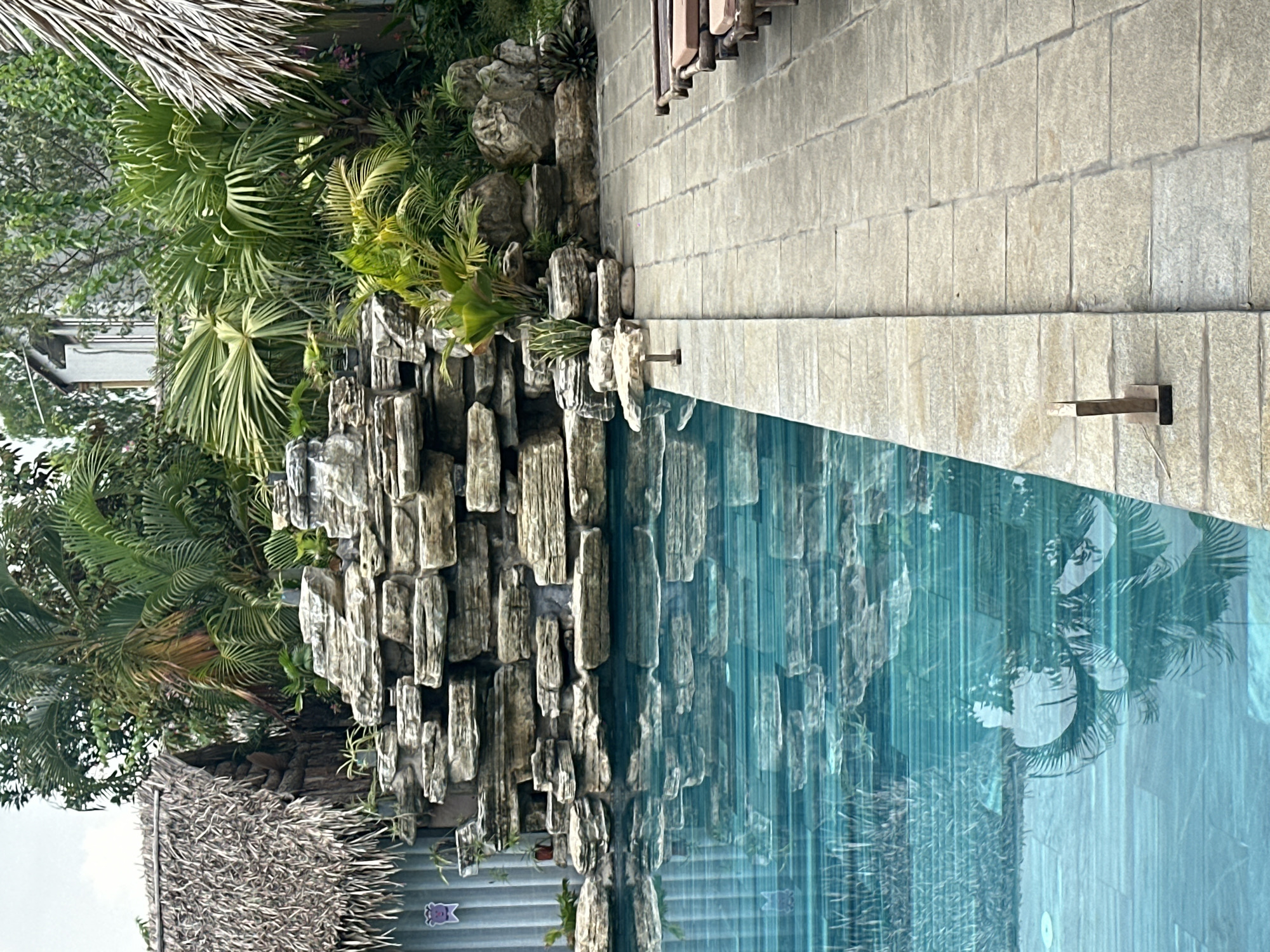 The image shows a swimming pool with a rock waterfall feature. The pool is bordered by a stone patio with lounge chairs. Lush tropical foliage, including palm trees, surrounds the pool area, with hints of thatched-roof structures in the background, suggesting a resort or hotel setting. There are no people visible in the image. The scene appears to be a serene outdoor recreational area.