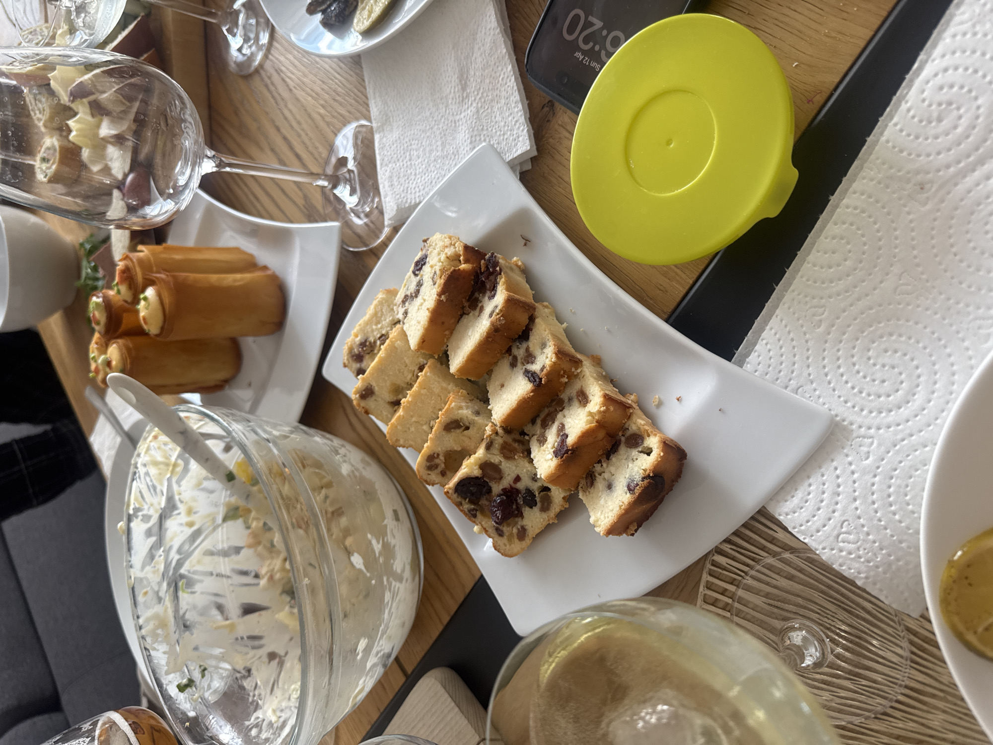 The image depicts a table scene indoors. The table is made of light brown wood. Visible on the table are plates of food, including sliced cake with raisins, rolled pastries, and a bowl of salad with a serving spoon. A yellow lid sits near a black smartphone displaying the time as 6:20 and the date as Sunday, April 12. There are also clear glasses, some containing liquids. Napkins are scattered on the table. A gray sofa is partly visible on the left. The location is likely a private residence in Warsaw, Poland.