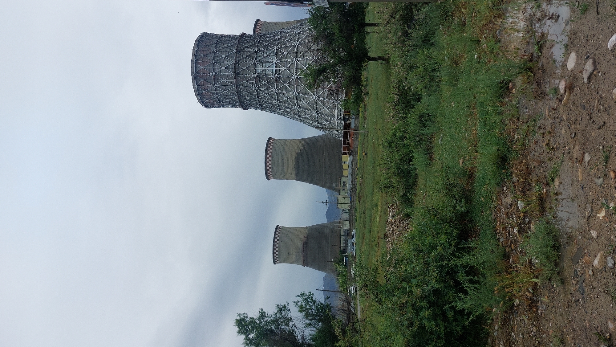 The image depicts three cooling towers, part of a power plant in the Taraz city administration, Kazakhstan. The closest tower is constructed with a distinctive lattice pattern at the top. The next two towers are made of concrete. A thin row of buildings and power lines are visible between the cooling towers and the distant mountain range. A dirt road covered in puddles runs along the right side of the frame. Green bushes and grass are abundant, lining the roadside. The sky is overcast.