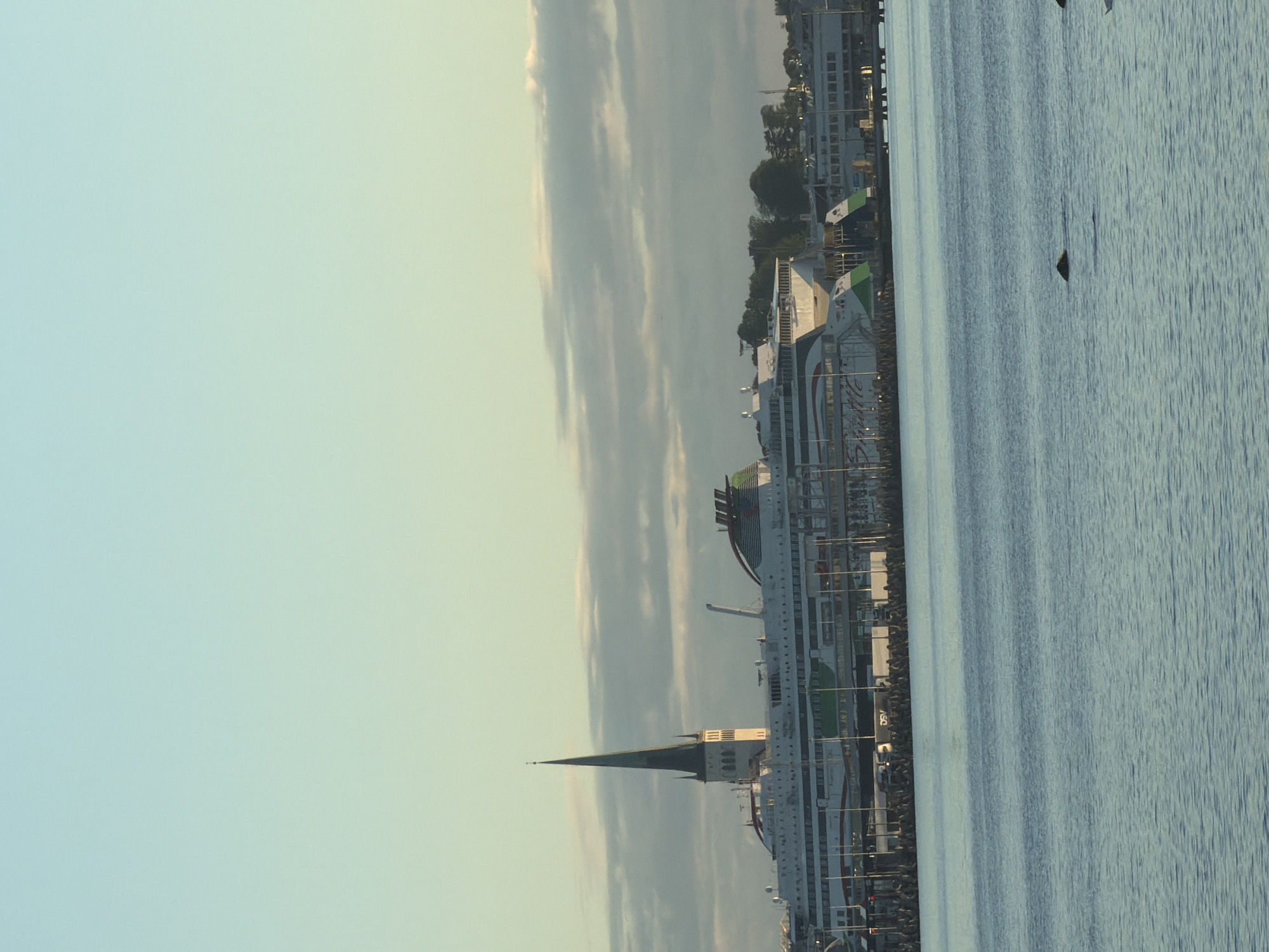 The image depicts a harbor scene in Tallinn, Estonia. Dominating the mid-ground is a large white ferry with green and red accents, docked at a pier. The ferry has multiple decks and is a significant presence in the port. To the left of the ferry, a tall, slender church spire with white and dark grey sections rises above the ship, suggesting its proximity to the harbor. In the background, other buildings and trees are visible, and further to the right, another, smaller ferry or ship is docked. The water in the foreground is calm and reflects the light from the sky. The sky itself is a soft gradient of pale blue and yellow, with scattered, elongated clouds. The overall impression is a tranquil morning or late afternoon in a bustling port city.