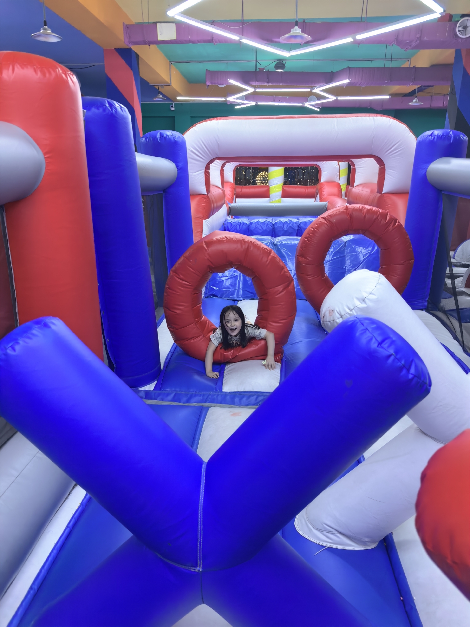 A young girl with dark hair, wearing a light-colored t-shirt, is partially visible inside a large, inflatable obstacle course. She is looking at the camera with an open-mouthed expression of excitement or surprise. The obstacle course is predominantly red, blue, and white, with large inflatable tubes forming various structures. The background shows the interior of a building with a colorful ceiling featuring fluorescent lighting and what appear to be air ducts. The floor of the obstacle course is a blue and white checkered pattern. The scene is an indoor playground or amusement center. This image was taken in Fergana City, Uzbekistan.