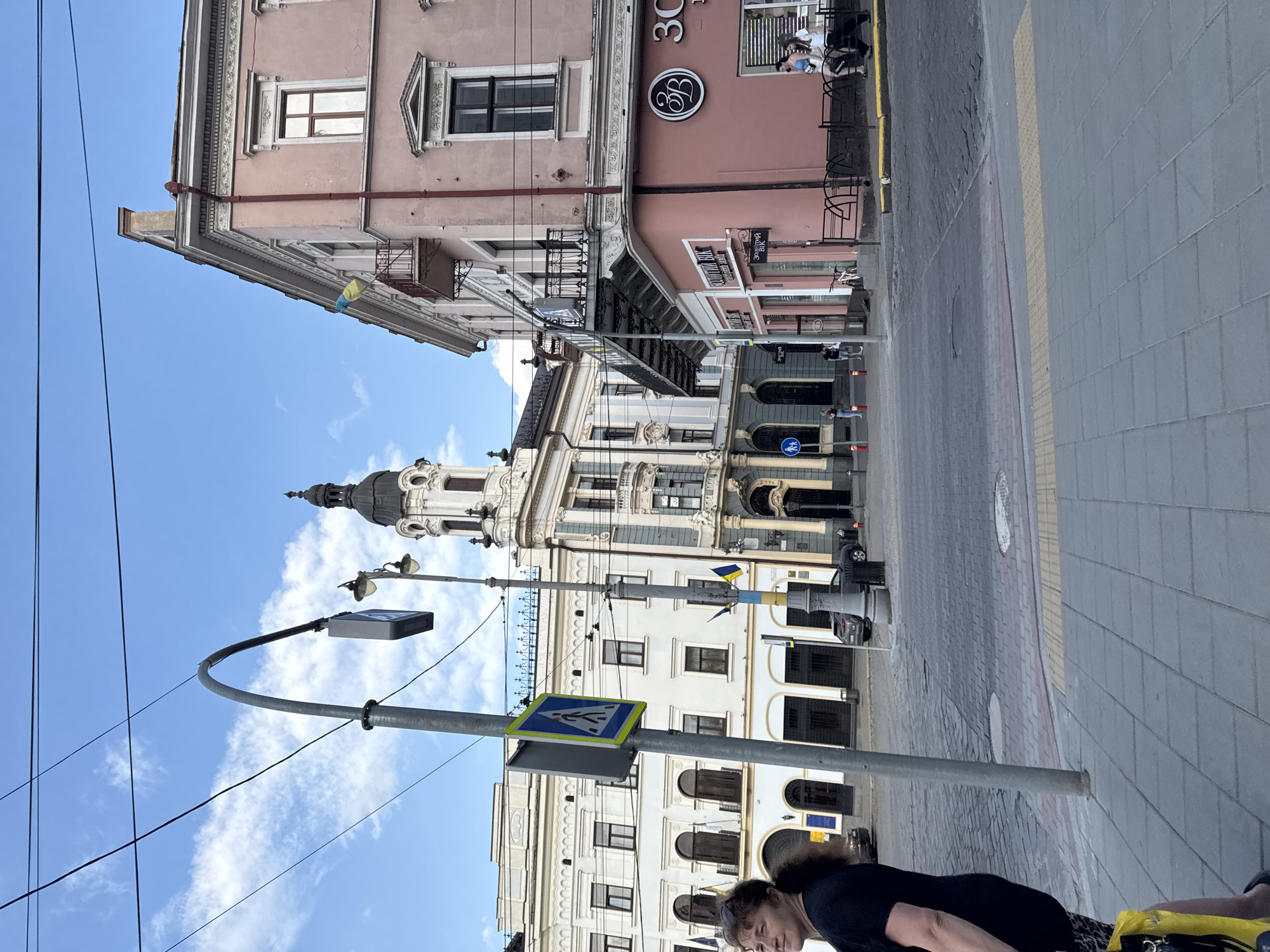 This image captures an outdoor urban scene in Chernivtsi, Ukraine, with a focus on the architecture and street life. The perspective is tilted, giving a dynamic view of the buildings and sky.
The subjects include several people, though their faces are not clearly discernible. One person in the foreground, in a black top and patterned skirt, appears to be walking or standing, with a yellow bag beside them. Other individuals are visible in the background, near storefronts and on the sidewalk.
The setting is a street lined with ornate, multi-story buildings exhibiting classic architectural styles. The buildings feature decorative facades, arched windows, and balconies. One building displays a prominent sign with a stylized "3C" logo and the text "3C Cafe", suggesting a commercial establishment. Another building has a dome-like structure at its peak. Ukrainian flags are visible, adding a national element to the scene.
Street furniture, including lampposts, a traffic sign for a pedestrian crossing, and bicycle racks, are present. The ground is paved with cobblestones and tiles. The time of day appears to be daytime, with bright sunlight casting shadows, indicating clear weather. The sky is blue with scattered white clouds.
The image depicts a moment of everyday life in a European city, with people moving through the street and interacting with the urban environment. The architectural details and the presence of national flags provide specific clues about the location and its cultural context.