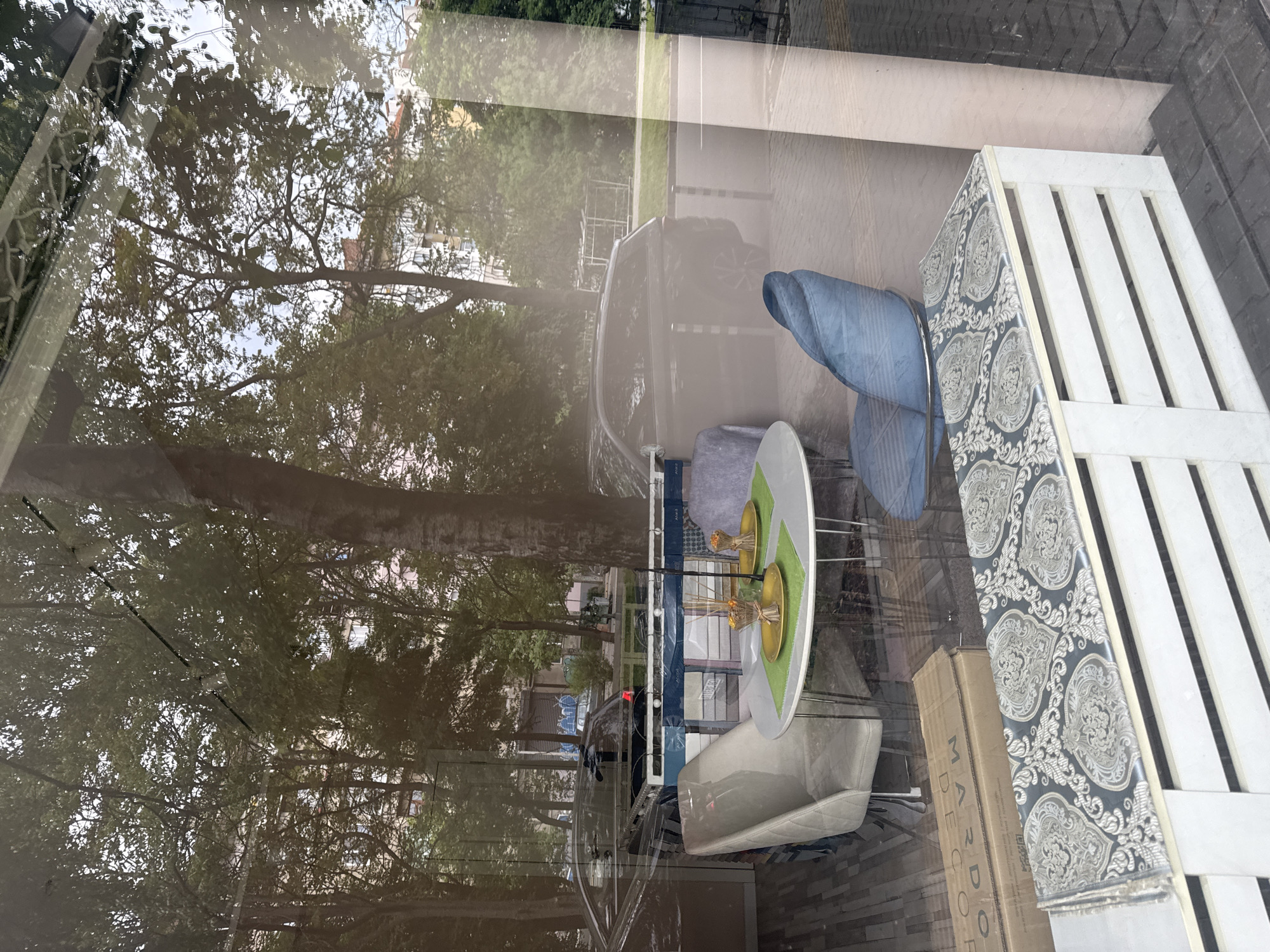 The image shows a storefront display reflecting a street scene. Inside, there's a table set with decorative items and chairs, as well as a section of shelving with patterned cushions and a box labeled "MARDO DECOR". The outside reflection shows a street with trees, a parked car, and buildings. Based on the environment, specifically the reflections and the store display contents, the location appears to be in a moderately affluent area of Sofia, Bulgaria.