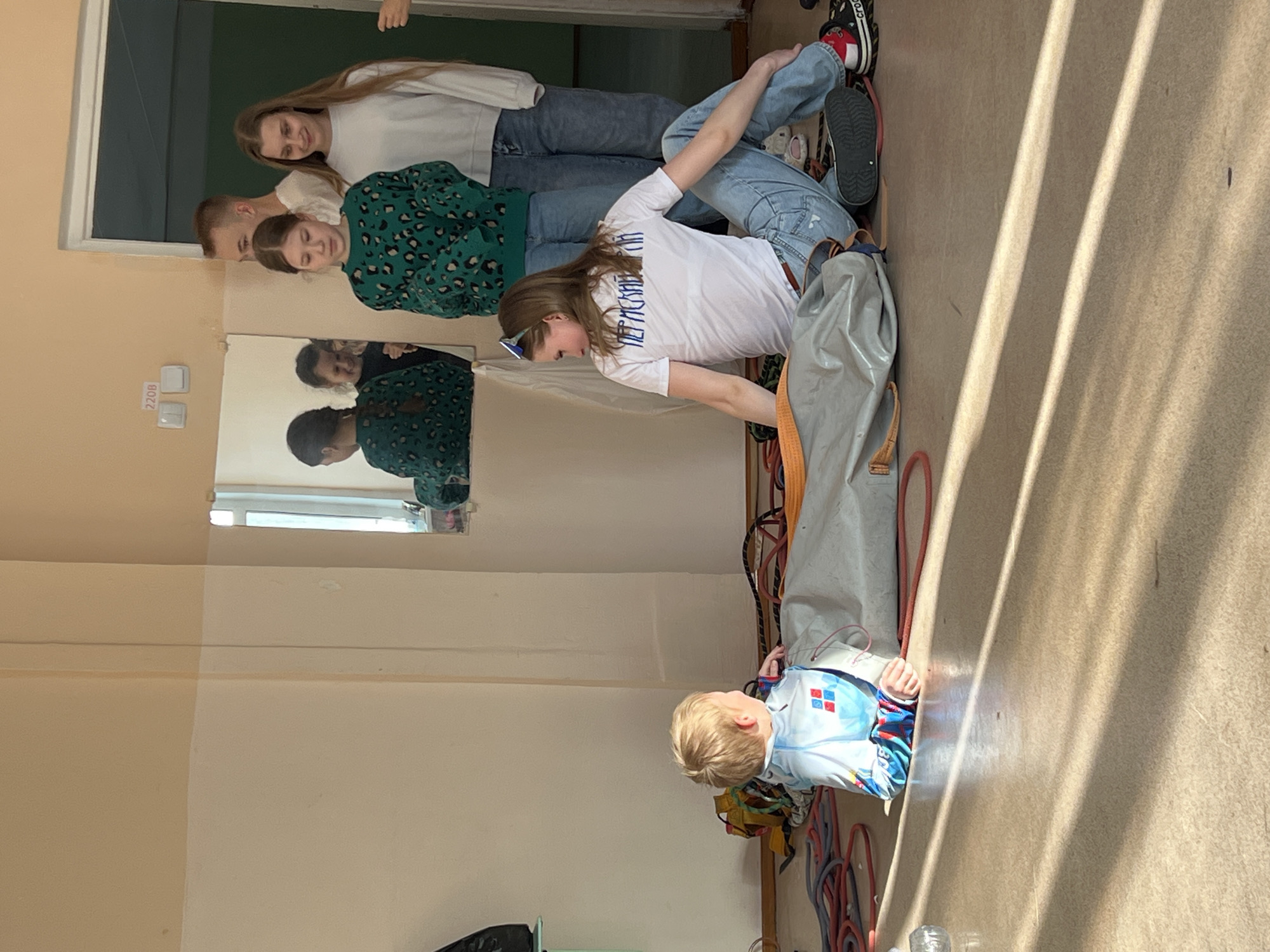 The image shows a group of four people standing in a room, with a child lying on the floor at their feet. The location is Berezniki, Russia, as indicated by the text on the woman's shirt.
From top to bottom, the group includes: a young woman in a white sweater and jeans, a male in a green leopard print sweater, a young girl in jeans and a white t-shirt with blue writing, and a blond child on the floor near a bundle of ropes. The child is wearing a light blue jacket. The three people standing near the top are lined up next to one another, and appear to be looking toward the camera. The room has light colored walls and a mirror on the wall. The area at the child's feet looks to contain climbing gear or ropes. There is a door visible at the top of the frame.