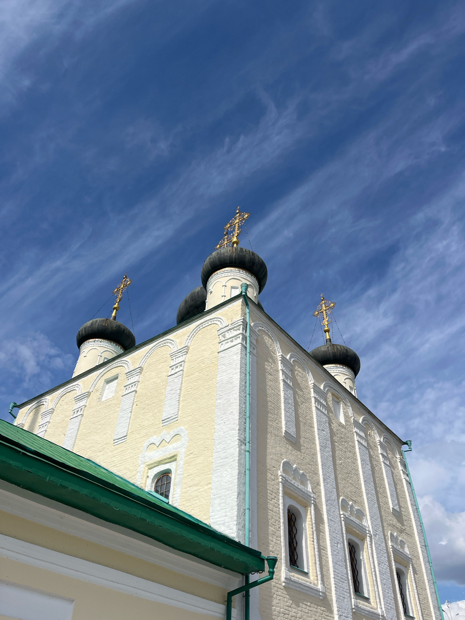 The image captures a low-angle view of the exterior of a historical church building, identified as the Church of the Assumption in Novaya Usman', Russia. The church features a pale yellow facade with white decorative elements and arched window frames. Several black domes topped with ornate gold crosses are visible against a vivid blue sky filled with wispy white clouds. A portion of a green roof runs along the bottom left of the frame, indicating it's part of a larger complex or an adjacent structure. The architectural style suggests an Orthodox Christian church, with multiple onion domes being a common characteristic. The lighting suggests it is daytime, likely with bright sunlight casting subtle shadows. No people are visible in the frame. The scene is a static architectural detail of a religious site.