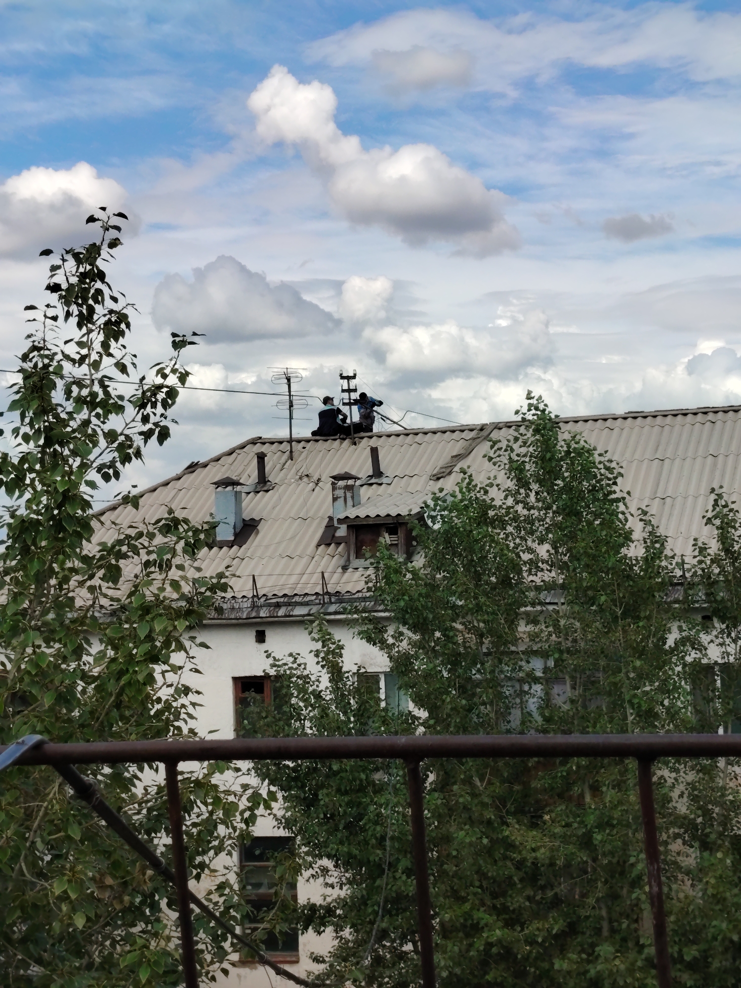 The image depicts a residential building's roof in Astana, Kazakhstan, under a cloudy sky. Two individuals are visible on the roof, seemingly engaged in some activity near antennae and electrical wires. The roof is covered with corrugated material and features several chimneys. Trees with green foliage flank the building, obscuring parts of its facade, which appears to be white. In the foreground, a dark-colored metal railing is partially visible. The overall scene suggests an urban residential setting.