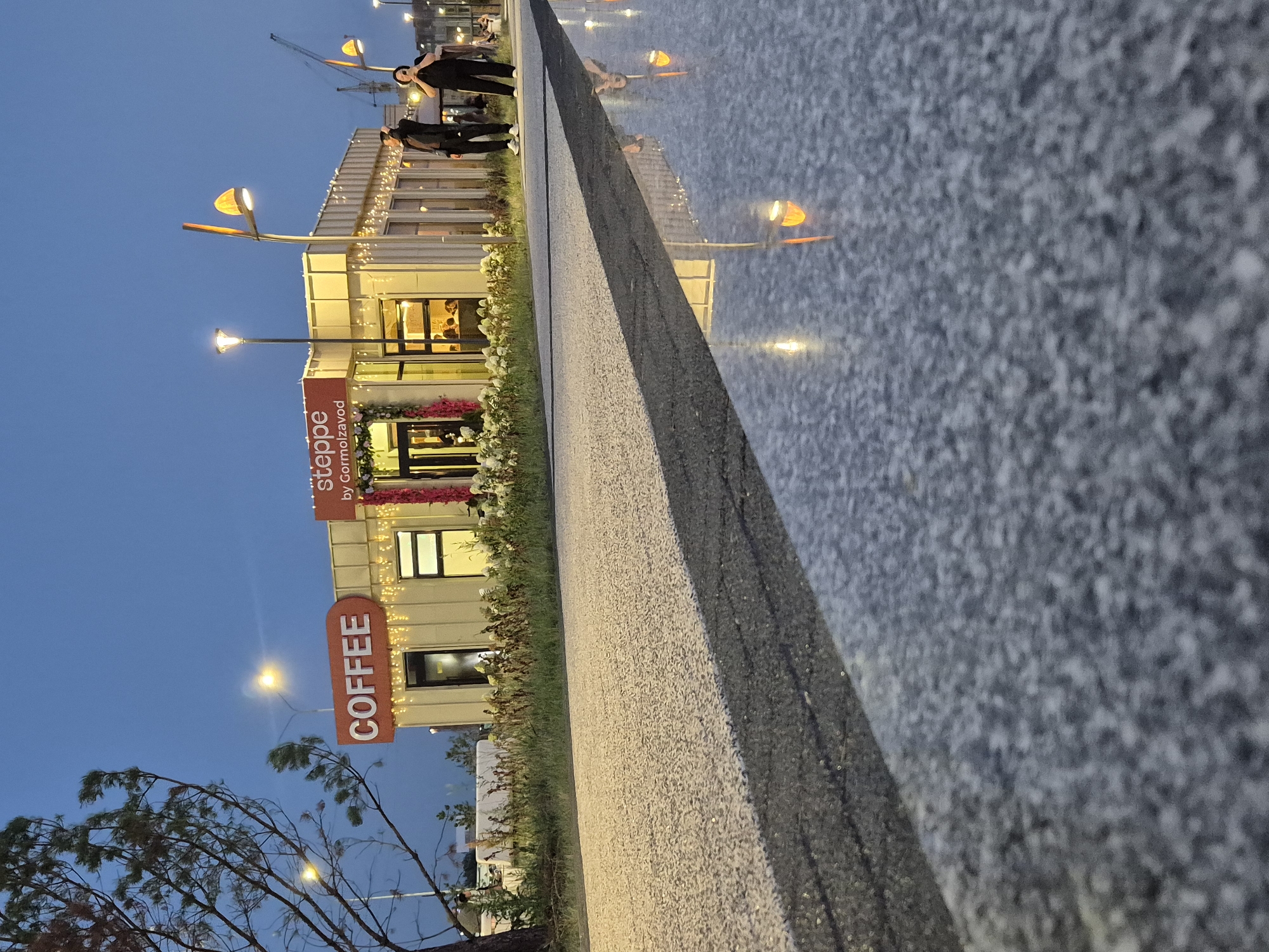 The image shows an outdoor scene at dusk, likely in Kokshetau, Kazakhstan. In the background, a modern building with large windows is illuminated by warm lights and string lights. A prominent orange sign reads "COFFEE" vertically, and another sign above it says "steppe by Gormol'zavod". People are visible inside the building and standing outside near the entrance, some appearing to be staff or patrons. The building is flanked by greenery and flowers.
In the foreground, a reflective surface, possibly a wet pavement or a shallow water feature, mirrors the lights from the building and surrounding streetlights. The texture of the reflective surface is granular and mottled. A dark, textured border runs diagonally across the frame, separating the reflective surface from a lighter, speckled walkway. A bare tree with wind-swept branches is visible on the left side of the image.
The sky is a deep blue, indicating the transition from day to night. The lighting is artificial and warm, creating a cozy atmosphere. The overall impression is that of a lively establishment, perhaps a café or restaurant, enjoying a soft evening. The presence of construction cranes in the far background suggests that the area might be undergoing development.