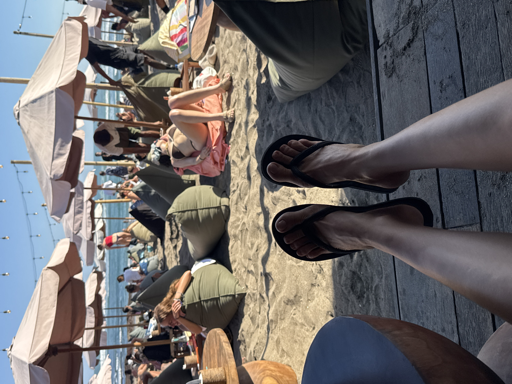 The image depicts a beachside scene in North Kuta, Indonesia, likely at a popular beach club. In the foreground, a pair of bare feet wearing black flip-flops are visible, suggesting the viewpoint of someone relaxing at the location. The background reveals a busy beach with numerous people scattered across the sand. Large, cream-colored umbrellas provide shade, and various beanbag chairs are set up for seating. The ocean is visible in the distance under a clear blue sky, with some individuals wading or swimming. People are engaged in various activities, including sunbathing, socializing, and preparing food or drinks. The overall atmosphere appears to be one of leisure and recreation.