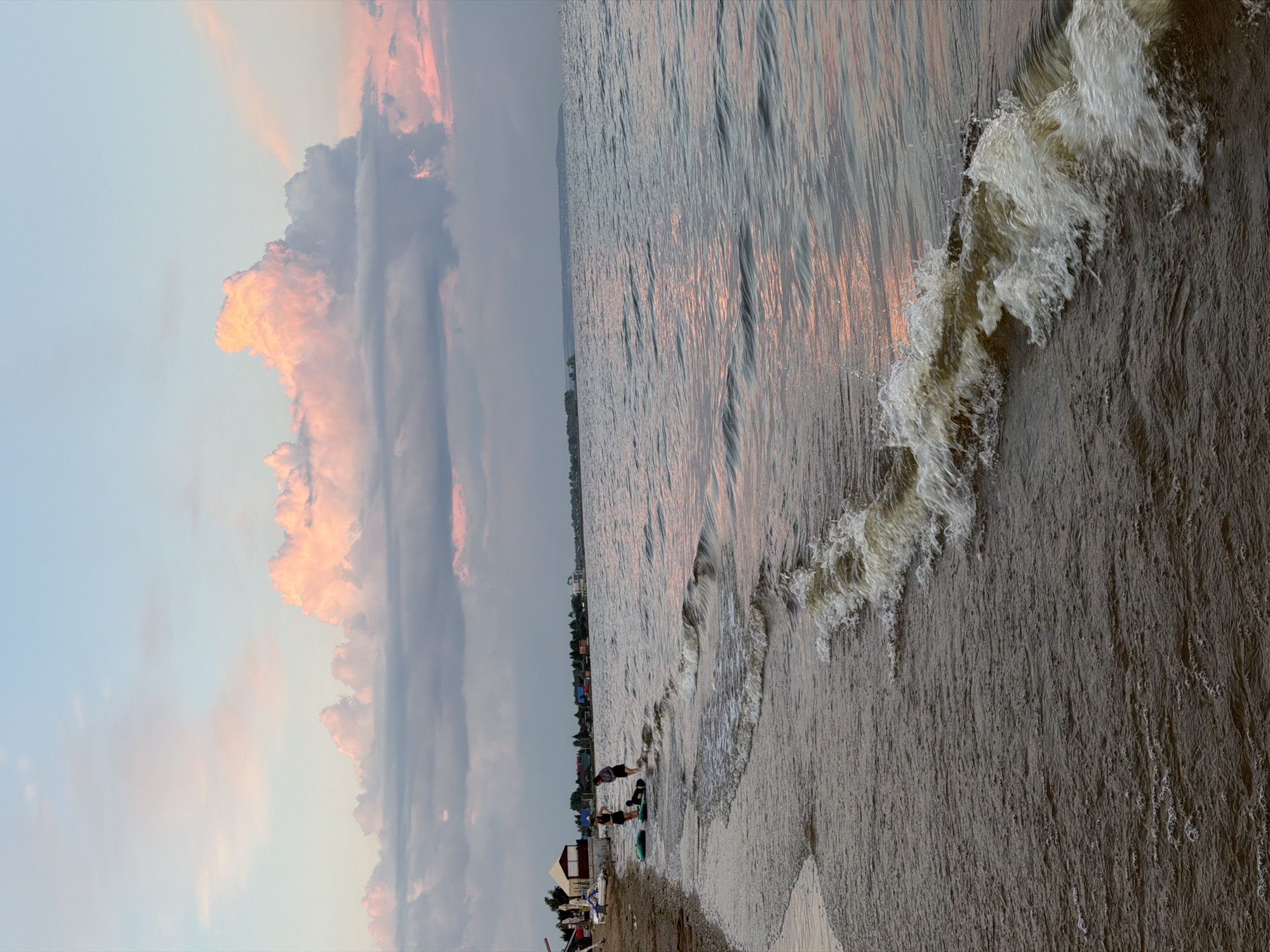 The image depicts a serene beach scene at sunset in Laishevo, Russia. The sky is a soft blend of blue and pink hues, with large, fluffy clouds tinged with the warm colors of the setting sun. A calm body of water, likely the Kama River, stretches across the frame, reflecting the sky's colors. Gentle waves lap at the sandy shore, where two individuals are present. One person, dressed in dark clothing, is standing near the water's edge, holding a wakeboard. Another person, also in dark clothing, is crouched next to a second wakeboard on the beach. In the background, a few buildings and trees are visible, hinting at the proximity of a town or resort. The overall atmosphere is peaceful and tranquil, capturing the beauty of a summer evening by the water.