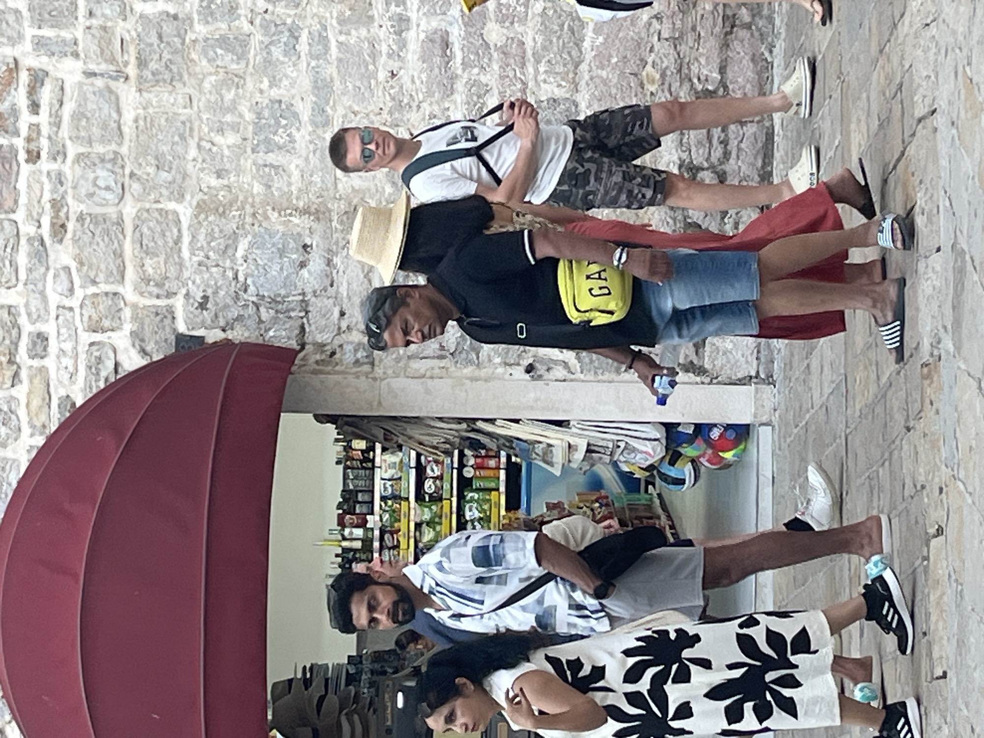 This is a medium-shot, eye-level, outdoor photograph of a group of people walking on a stone-paved sidewalk in front of a store. The store appears to be a souvenir shop, with shelves stocked with various goods visible through an open doorway. A large, dark red awning extends over the storefront. The architecture suggests a European setting, consistent with Budva, Montenegro.
In the foreground, from left to right:
A woman wearing a white sleeveless dress with a bold black floral pattern walks towards the left. She has long, dark hair and is looking to her left.
A man wearing a white short-sleeved button-up shirt with a blue geometric pattern, grey shorts, and a black crossbody bag walks towards the right. He has dark hair and a beard and is looking forward.
A man wearing a black polo shirt, denim shorts, and a bright yellow fanny pack is walking towards the right. He has short grey hair and is holding a clear plastic water bottle in his left hand.
A person wearing a wide-brimmed straw hat and a rust-colored dress is walking behind the man in the black polo shirt. Only the hat and the side of the dress are clearly visible.
A man wearing a white t-shirt, camouflage shorts, and sunglasses is walking towards the right. He has short blonde hair and a backpack strapped to his shoulders. He is looking towards the viewer.
In the background, behind the people, shelves in the store are visible, filled with bottles, packaged goods, and other items. The wall behind the people is constructed of rough, grey stone blocks. The sidewalk is made of square-shaped paving stones. The lighting suggests it is daytime.