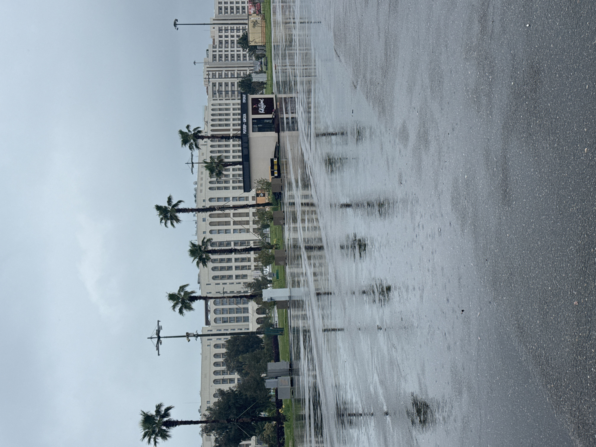 The image depicts a wet, paved area, likely a road or sidewalk, reflecting the surrounding urban landscape. A row of tall, white buildings lines the left side of the image, interspersed with palm trees. There are light poles positioned along the row of buildings. A building with signage in a Cyrillic script is visible midway down the row. The sky is overcast and grey, suggesting a rainy day. The overall impression is one of a modern urban area after rainfall. Based on information from the web, Sirius is a city in Russia and the Cyrillic script on the building and the modern architecture of the buildings could indicate that this image was taken in Sirius, Russia.