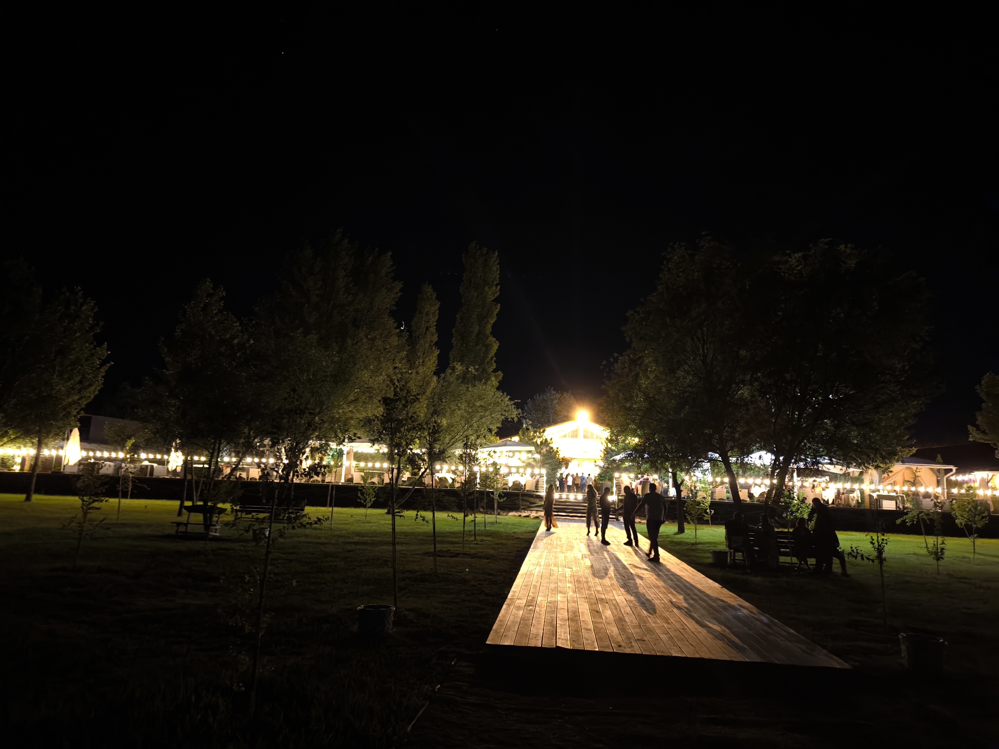 In an outdoor setting at night, a wooden pathway with pronounced plank lines extends diagonally across the foreground, illuminated by overhead lights. Several individuals, silhouetted against the bright pathway, are visible standing and walking. To the left and rear of the pathway, a grassy area is dotted with young trees and benches. Beyond the lawn, a building with extensive string lights is illuminated, suggesting a restaurant or event venue. Trees with dense foliage are scattered throughout the scene, with their branches silhouetted against the dark night sky. The overall impression is of an outdoor gathering or dining area at night, with a focus on the illuminated pathway and the lively atmosphere of the venue.