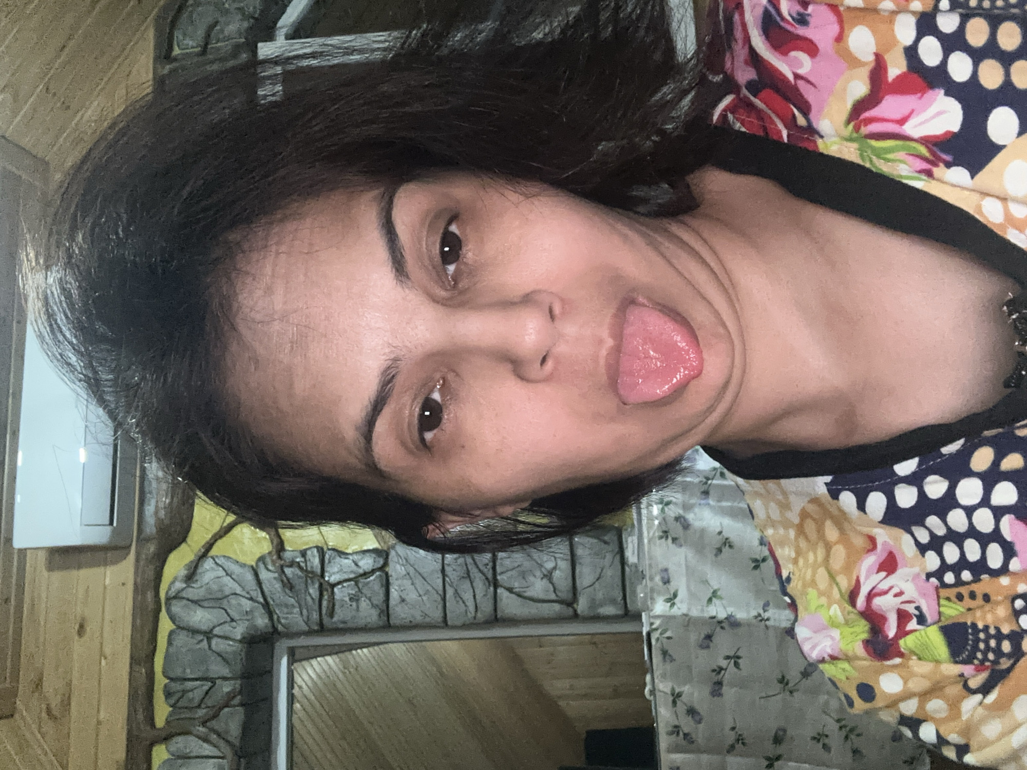 A woman is sticking her tongue out at the camera in a selfie. She has dark hair and is wearing a colorful, patterned shirt. The background appears to be indoors, with wooden paneling on the ceiling and a stone-like wall decoration. An air conditioning unit is visible on the wall. The location is not specified as Kalininskaya, Russia in the image.