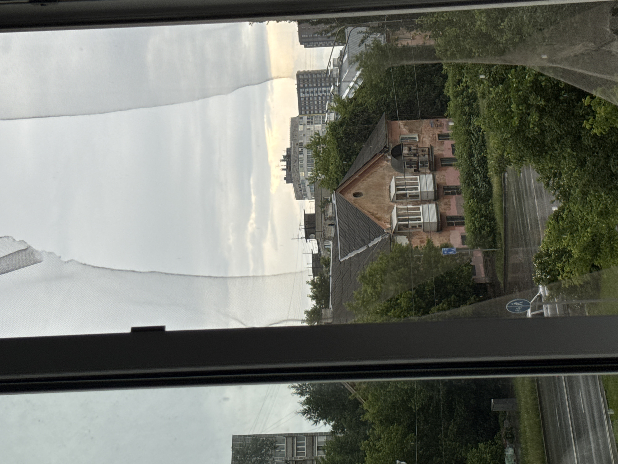 The image presents a view from an indoor window looking out onto an urban landscape, likely in Perm, Russia, under an overcast sky. The dark frame of the window occupies the foreground, along with a torn white curtain or mesh partially visible on the left side. Reflections of a building and trees can be seen on the window glass, particularly in the lower portion.
Beyond the window, a wet road or street, suggesting recent rainfall, cuts through the scene. It is lined with numerous lush green trees. A prominent, older-style building with a reddish-pink facade, a dark sloped roof, and multiple windows (some with white frames, others with metal covers) stands centrally. It features a small circular vent or window on its front and a small balcony. In the background, several taller, more modern multi-story buildings, ranging from dark to light gray, rise above the treeline against the cloudy sky.
The sky is heavily overcast, though patches of lighter clouds indicate it is daytime, possibly morning or late afternoon. There is no visible human activity. A circular blue road sign with white pictograms of a pedestrian and a bicycle is clearly visible near the street, indicating a shared path. The mix of historical and contemporary architecture, combined with the wet streets and overcast conditions, paints a quiet, typical city scene.