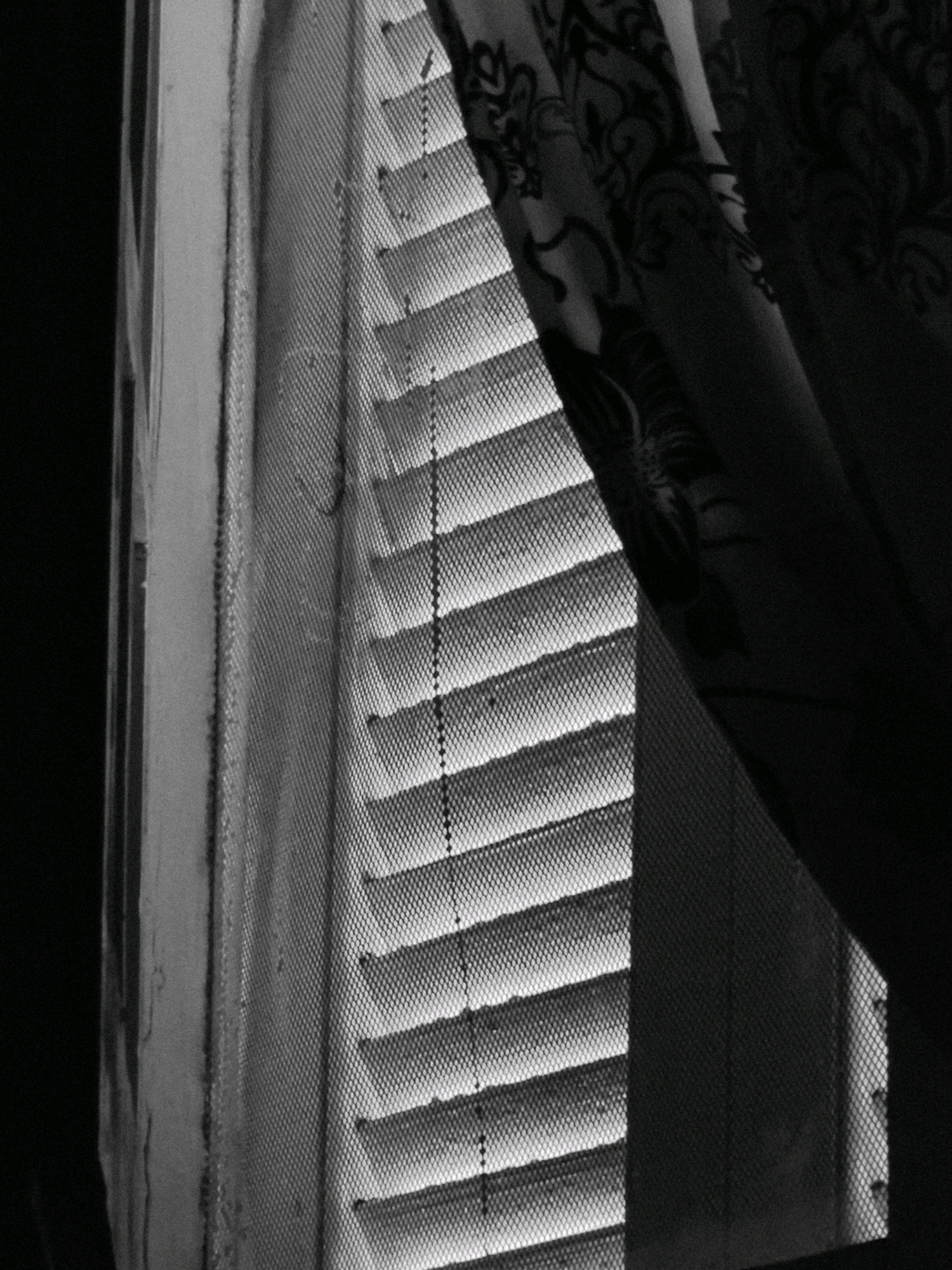 The image displays a close-up, black and white view of a window with Venetian blinds. The blinds are partially open, allowing light to filter through in horizontal stripes. A patterned curtain is drawn to the right side of the window, partially obscuring the view. The window frame appears to be made of weathered material, possibly wood or stucco, with some texture visible. The overall impression is one of indoor ambiance, with the play of light and shadow suggesting a dimly lit interior. The scene is devoid of people, animals, or any overt actions. The location in Al Khusus, Egypt, is not discernible from the visual elements alone.