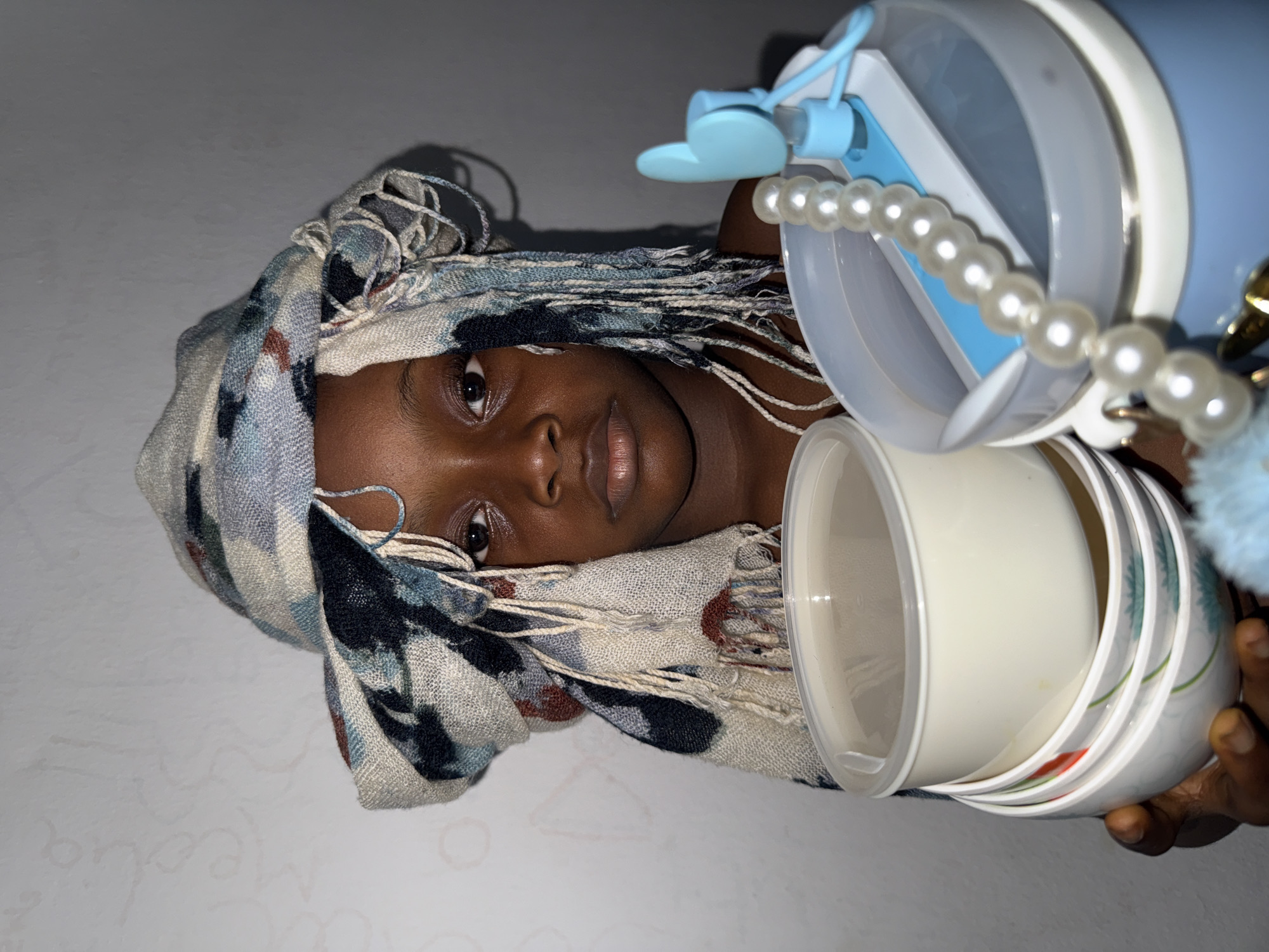 A close-up indoor shot shows a woman with dark skin wearing a patterned headscarf, lying on a white surface. The headscarf is a mix of light blue, gray, and black, with a fringed edge that falls over part of her face. Next to her head is a stack of white bowls or containers, held in place by the woman's hand. A pearl necklace drapes over these containers. In the background, there's a blue and white water bottle. The location is likely a residential setting, but specific details to identify it as being in Ado, Nigeria, are absent.