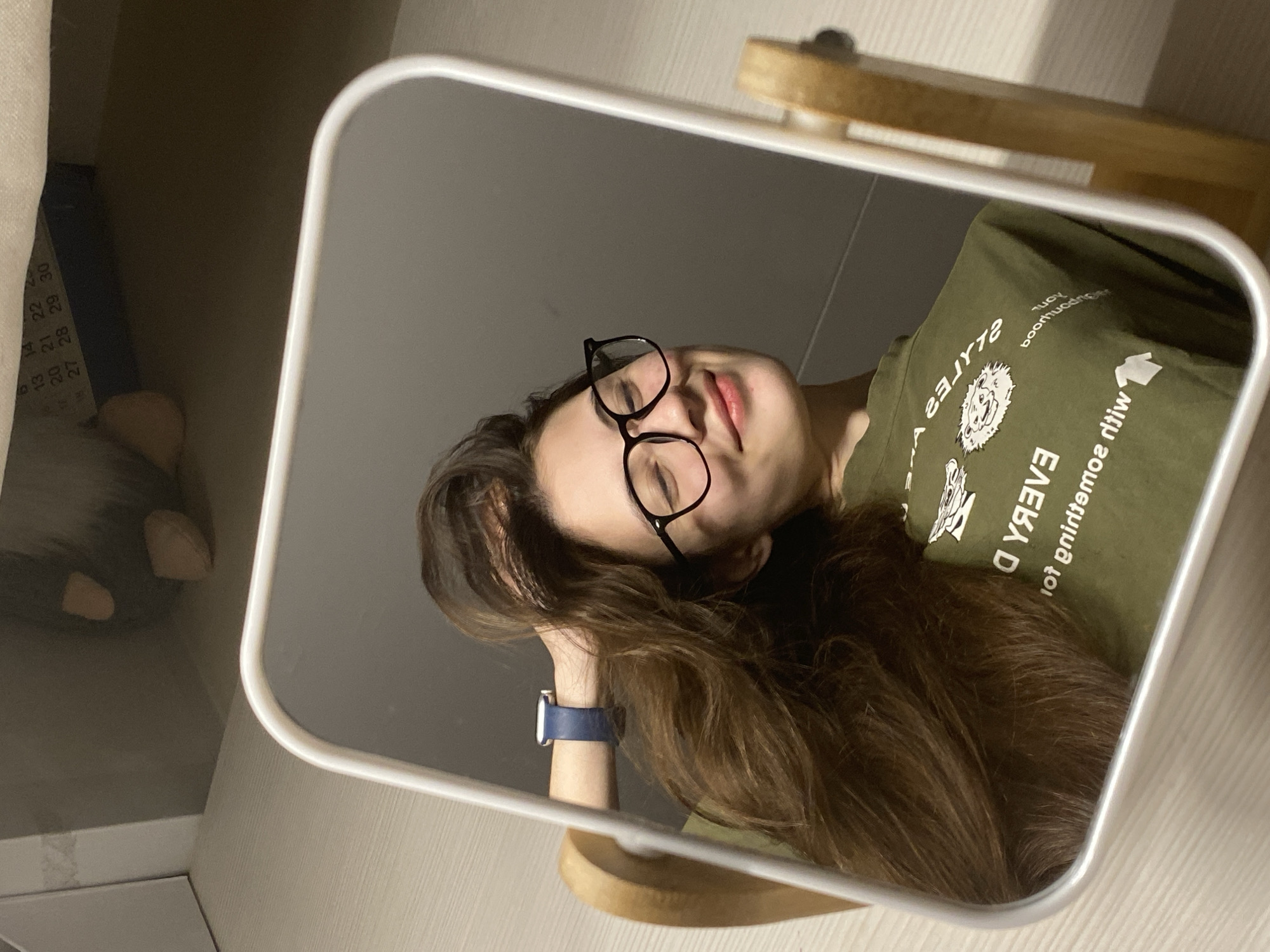 An image, captured at an unusual clockwise rotation, displays a mirror selfie of a young woman in an indoor setting. The woman has long, wavy dark brown hair and wears black-framed glasses. She is dressed in a dark green t-shirt featuring a white graphic of a lion's head and text, which appears reversed in the mirror but reads "EVERY DAY is for something new with you." A dark blue watch is visible on her left wrist. Her eyes are closed, and a gentle smile suggests a relaxed or peaceful mood. Her left hand rests casually on her hair.
The rectangular mirror has a light-colored frame and stands on a wooden base. The background reflected in the mirror is dimly lit and indistinct. Outside the mirror, on the left, a light-colored fabric and a partial calendar or planner are visible, showing numbers like "13, 14, 20, 21, 22, 27, 28, 29, 30". A blurry, dark object, possibly a plush item, is also in the background. The scene is likely a personal room in Votkinsk, Russia.