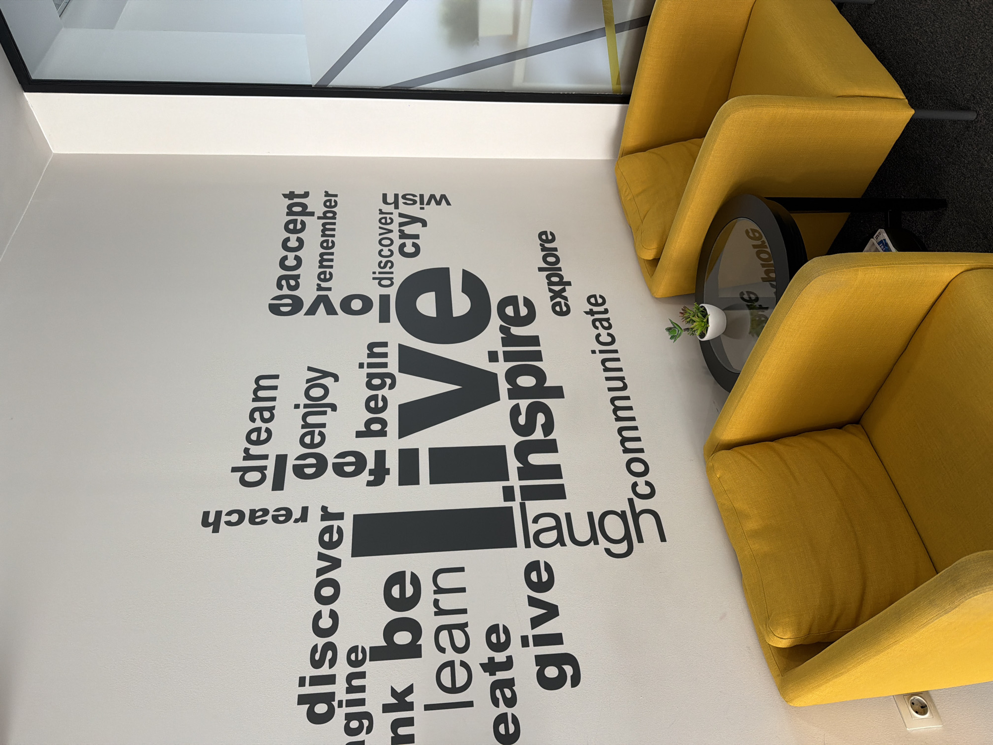 This image shows a corner of a room with two yellow armchairs placed next to a small round table with a plant on it. The wall is white with black lettering forming a collage of inspirational words like "dream," "live," "inspire," "discover," "learn," and others. The floor is gray. Based on the style and decor, it might be located in a modern office or communal space.
However, there is no information available about the location in Sofia, Bulgaria, in the image.