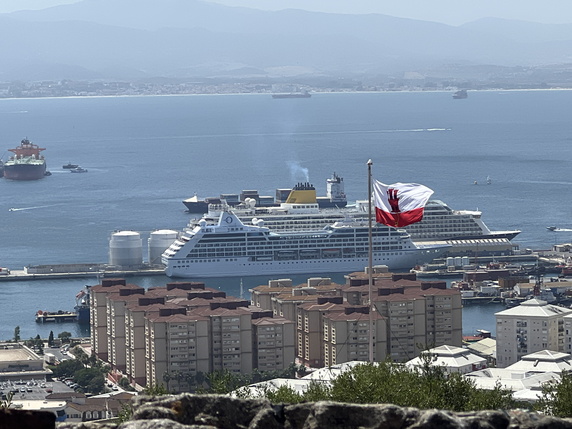The image depicts a harbor scene in Gibraltar, Spain, with a large cruise ship docked in the background. In the foreground, a flag of Gibraltar is prominently displayed on a flagpole, its red and white colors stark against the blue sky. Several residential buildings with red-tiled roofs are visible, overlooking the harbor. Other ships, including cargo vessels and smaller boats, are scattered throughout the water. The background shows a distant coastline with a range of hills or mountains. The scene appears to be a sunny day, with clear skies and calm waters.