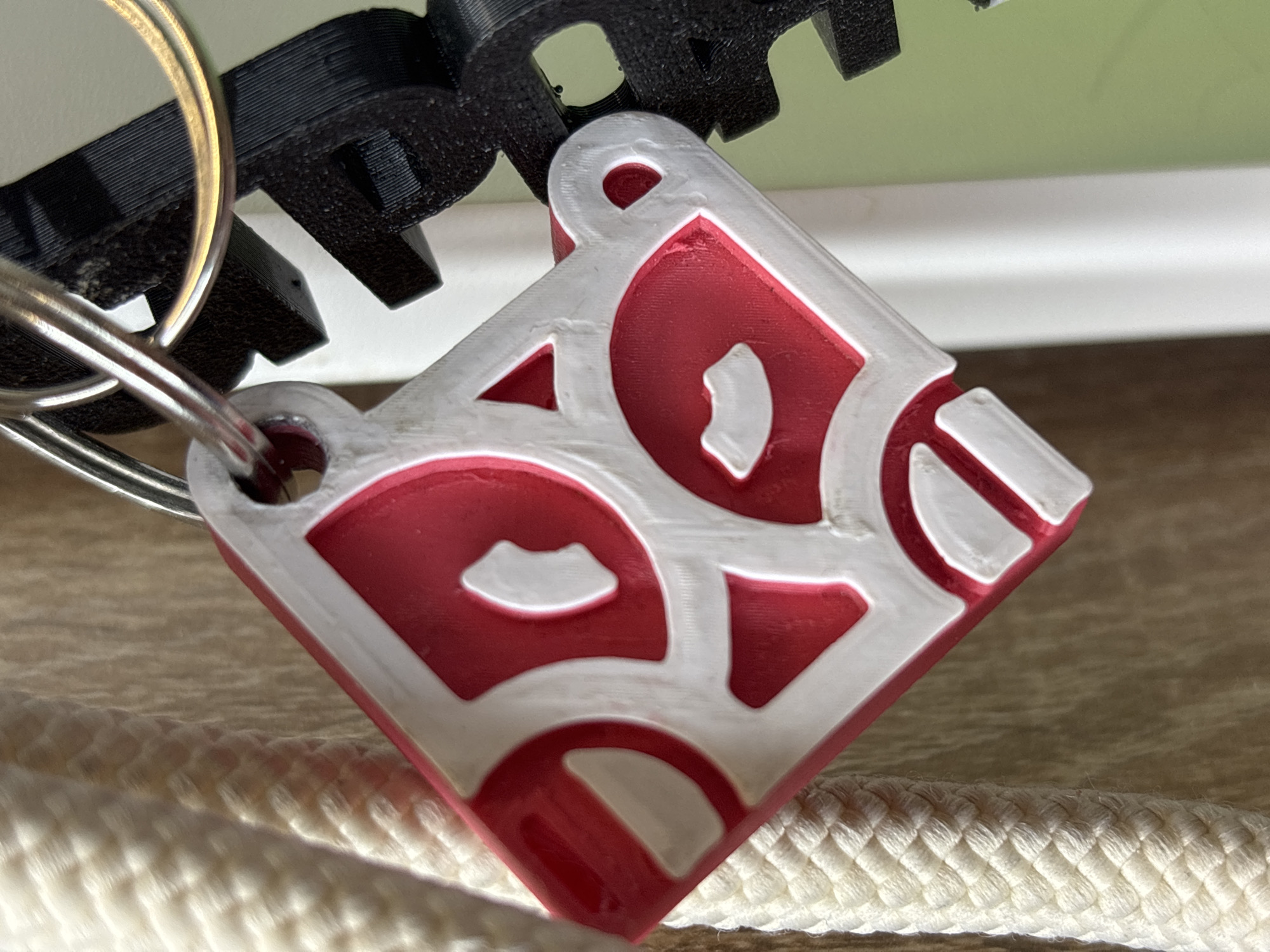 A close-up shot depicts a square keychain with the letters "PERM" in a stylized font. The letters are colored in red with white outlines. The keychain is hanging from a metal ring, which is connected to a black, 3D-printed object with gear-like protrusions. The keychain rests on a white, braided cord. The background features a pale green surface above a white, horizontal plane. The city of Perm, Russia is inferred from the lettering on the keychain.