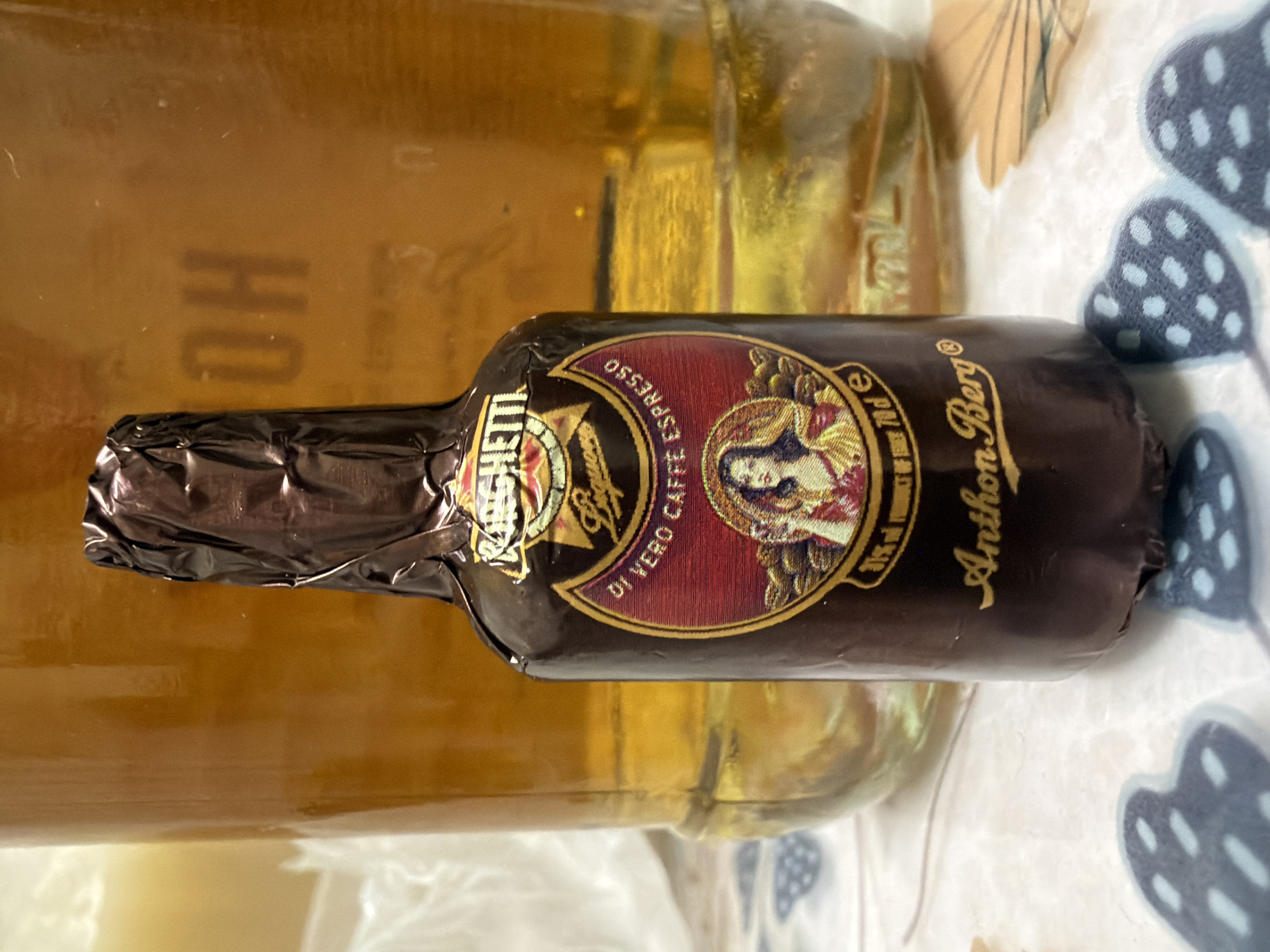 This is a close-up, eye-level, indoor shot of a small bottle wrapped in dark brown foil, similar to a miniature liquor bottle. The bottle stands upright in the foreground, slightly off-center to the right. It has a prominent label with a circular design featuring a woman's portrait and text. The text on the label includes "RONCHETTI," "Baguio," "DI VERO CAFFÈ ESPRESSO," and "Anthon Berg." Below the circular design, there is smaller text that appears to be a percentage and "70 cl e."
Behind the small bottle, and significantly larger, is a glass bottle or container filled with a golden amber liquid. This larger container is mostly out of focus but shows some faint markings on its surface, including what looks like "OH."
The surface on which the bottles are placed is a light-colored cloth with a patterned design of dark blue hearts filled with light blue dots. In the background, there are also some light brown, fan-shaped decorative elements scattered on the cloth. The lighting appears to be artificial and warm, casting soft shadows.
There are no people visible in the image. The scene is static, with no apparent action or interaction. The composition focuses on the products, suggesting a still-life arrangement. The image does not provide any specific information about its location in the city of Kyiv, Ukraine.