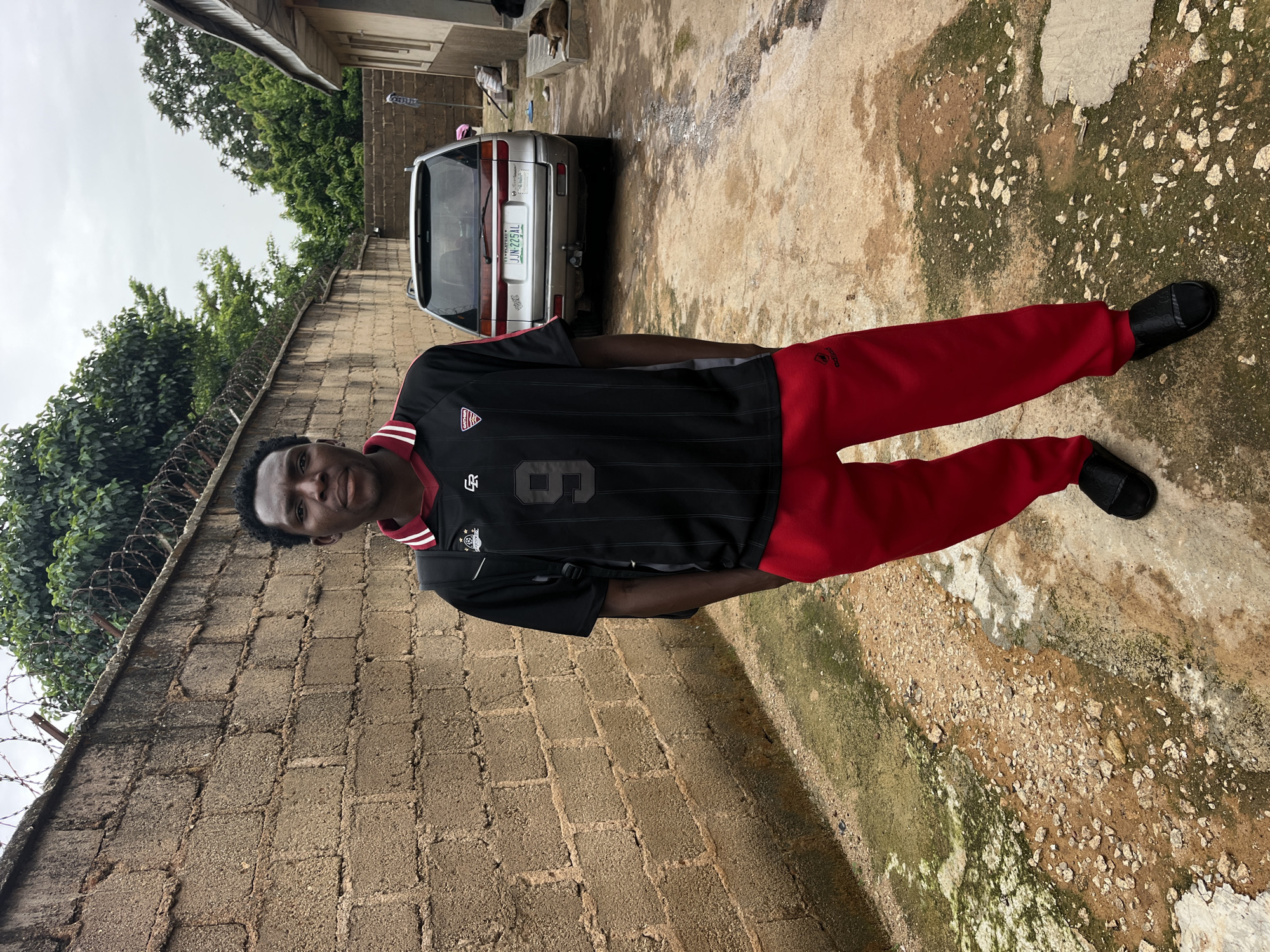 A young Black male stands in front of a brick wall, wearing a black and red soccer jersey with the number 9 on it, and red track pants. He has short, black hair and is looking directly at the camera. He is wearing black slides. A silver SUV is parked behind him. The ground is a mix of dirt, gravel, and concrete. There is barbed wire on top of the brick wall, and trees are visible in the background. The scene appears to be an outdoor courtyard or alleyway.