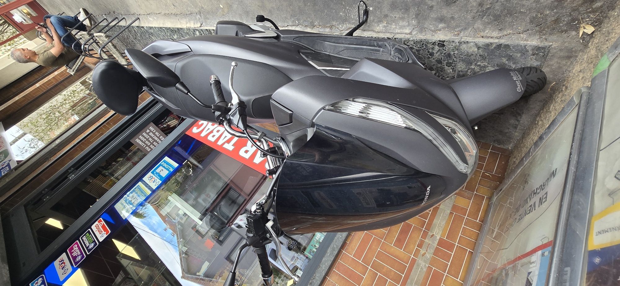 In an outdoor urban setting, a dark gray scooter is parked on a sidewalk next to a brick-faced building. The scooter's front is angled towards the viewer, with its headlights and handlebars clearly visible. A large reflective surface, possibly a store window, is in the background, reflecting the sky, trees, and buildings. Several logos and signs are visible on the store window, including "ALLIKO", "PARIONS SPORT", and "amigo". A red banner with white lettering "BAR TABAC" is diagonally placed across the window. To the right of the scooter, a man with gray hair is seated on a black outdoor chair, holding a smartphone. He is wearing a green t-shirt and blue jeans. The ground is paved with concrete and some debris is scattered around. The lighting suggests it is daytime.