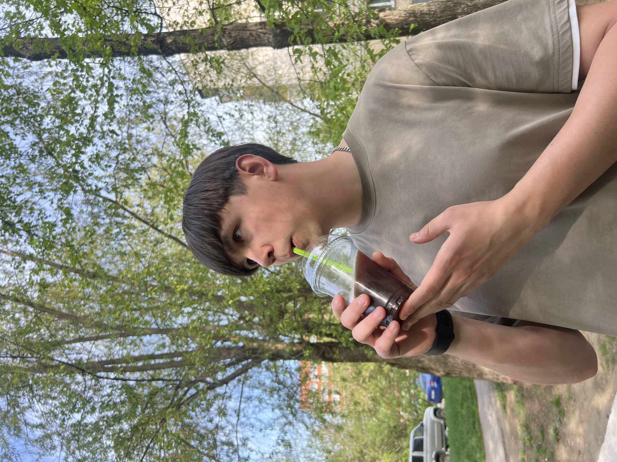 A medium shot depicts a young man outdoors, drinking from a transparent plastic cup with a green straw. He wears a beige t-shirt and a black wristband and stands against a backdrop of trees with fresh green leaves and a partly visible building. The location appears to be in the city of Chaikovsky, Russia. The man is holding the cup with both hands, focusing on drinking from the straw. The surroundings include a glimpse of a road, a vehicle, and a mix of urban and natural elements.