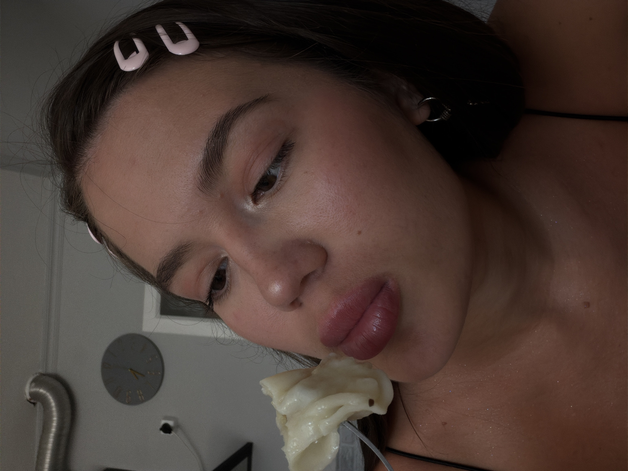 This media file is a close-up, portrait-style photograph of a young woman in what appears to be an indoor setting. The woman has dark hair, which is held back from her face with two light pink U-shaped hair clips. She is wearing a black top, and a subtle shimmer is visible on her collarbone and neck.
The woman is looking down and to the side, with her lips slightly parted as she holds a fork with food to her mouth. The food appears to be light in color and somewhat amorphous in shape. Her makeup includes blush on her cheeks, giving her skin a healthy glow. A small silver hoop earring is visible in her right ear.
In the background, a gray wall is visible with a round clock on it. The clock has gold numerals and hands, indicating a time around 1:45. To the left of the clock, a metallic silver pipe or duct is partially visible. The lighting suggests it might be evening or a dimly lit indoor space, as there are some shadows and the overall illumination is soft. No specific weather conditions are discernible. The image conveys a sense of casual intimacy, possibly capturing a moment of eating or posing.