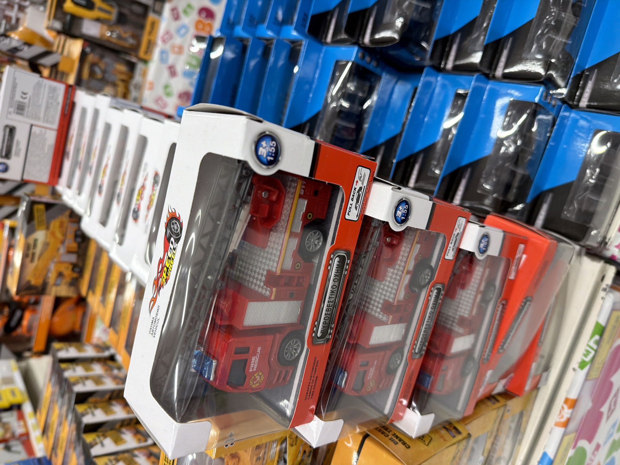 The image displays a close-up view of toy fire trucks packaged for sale on shelves within a retail store. The fire trucks are red and appear to be made of plastic and metal, with details like ladders and emergency lights visible. They are housed in transparent plastic packaging with red cardboard bases and white tops. Text on the packaging includes "FIRE RESCUE" and what appears to be Greek writing. Adjacent to the fire trucks, other toy vehicles, primarily construction trucks, are visible in their original packaging, featuring orange and yellow hues. The background shows rows of merchandise on store shelves, indicating a commercial environment. The scene is well-lit, consistent with indoor retail lighting. The focus is on the product display, with no people or identifiable landmarks in the immediate vicinity.