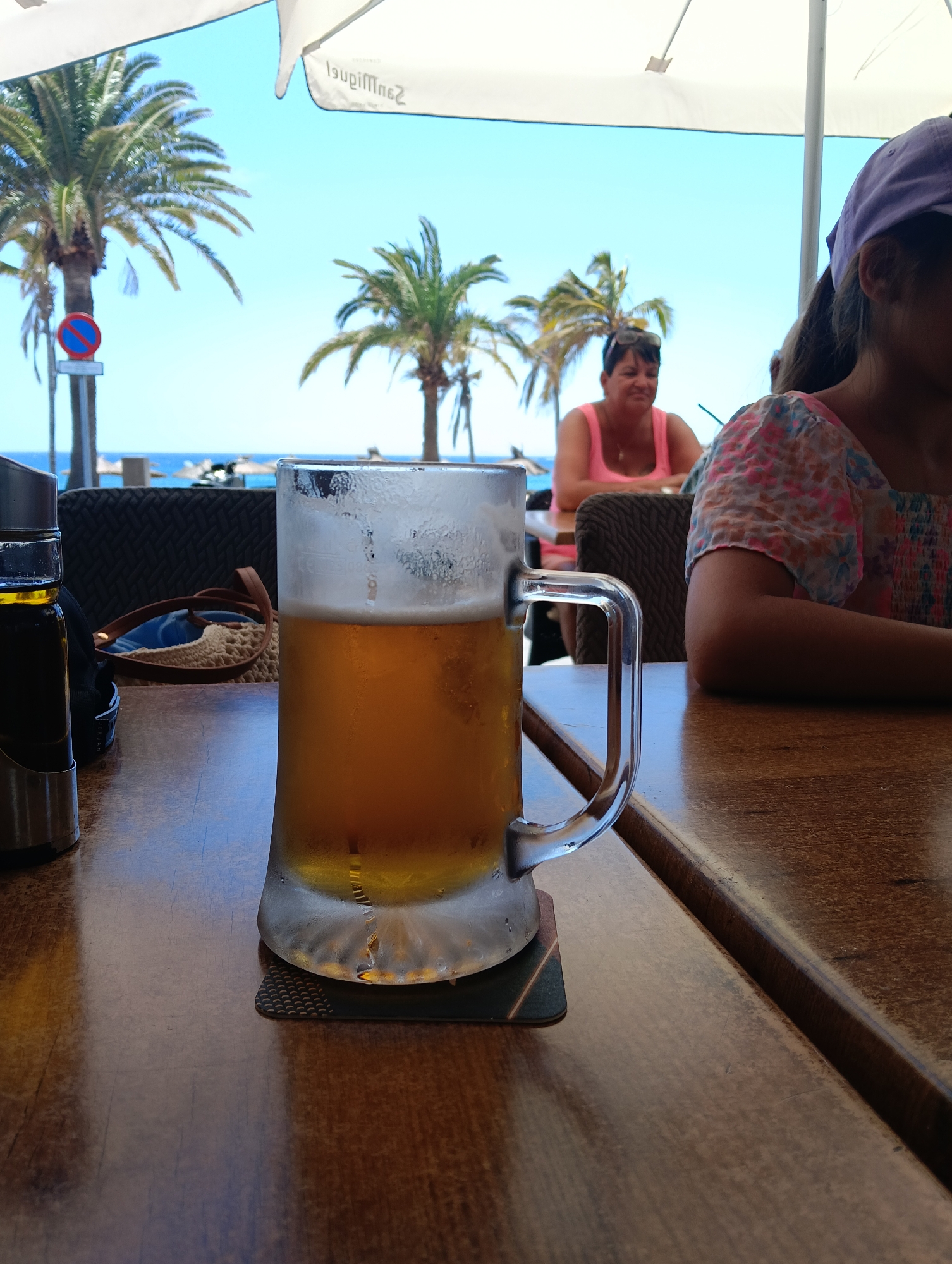 This vibrant image depicts an outdoor dining or bar setting, likely a beachside establishment in Puerto Del Carmen, Spain. The time of day is bright and sunny, suggested by the clear blue sky and well-lit scene.
In the immediate foreground, a large, frosted glass mug filled with golden beer rests on a dark coaster on a wooden table. Condiment bottles in a metal holder are visible to the left of the beer. The table surface is a warm, polished brown.
In the midground, several people are present. To the right, a person, likely a young female, is partially visible from the side, wearing a colorful floral top and a purple baseball cap. Further to the left and slightly behind, a woman with dark hair, wearing a bright pink tank top, is seated at another table, looking towards the right with a soft, content smile. On a table in the mid-left, sunglasses and a straw bag are casually placed.
The background showcases a beautiful coastal landscape. Tall palm trees punctuate the scene against a brilliant, cloudless blue sky. Beyond the trees, the sparkling blue ocean is visible, with distant beach umbrellas dotting the shore. A "no stopping or parking" sign (a red circle with a blue center and a red diagonal line) is discernible near one of the palm trees on the left. Overhead, a large white umbrella provides shade, and the reversed text "San Miguel" is clearly visible on its underside.
The overall scene captures a relaxed, leisurely vacation atmosphere, with people enjoying drinks by the sea.