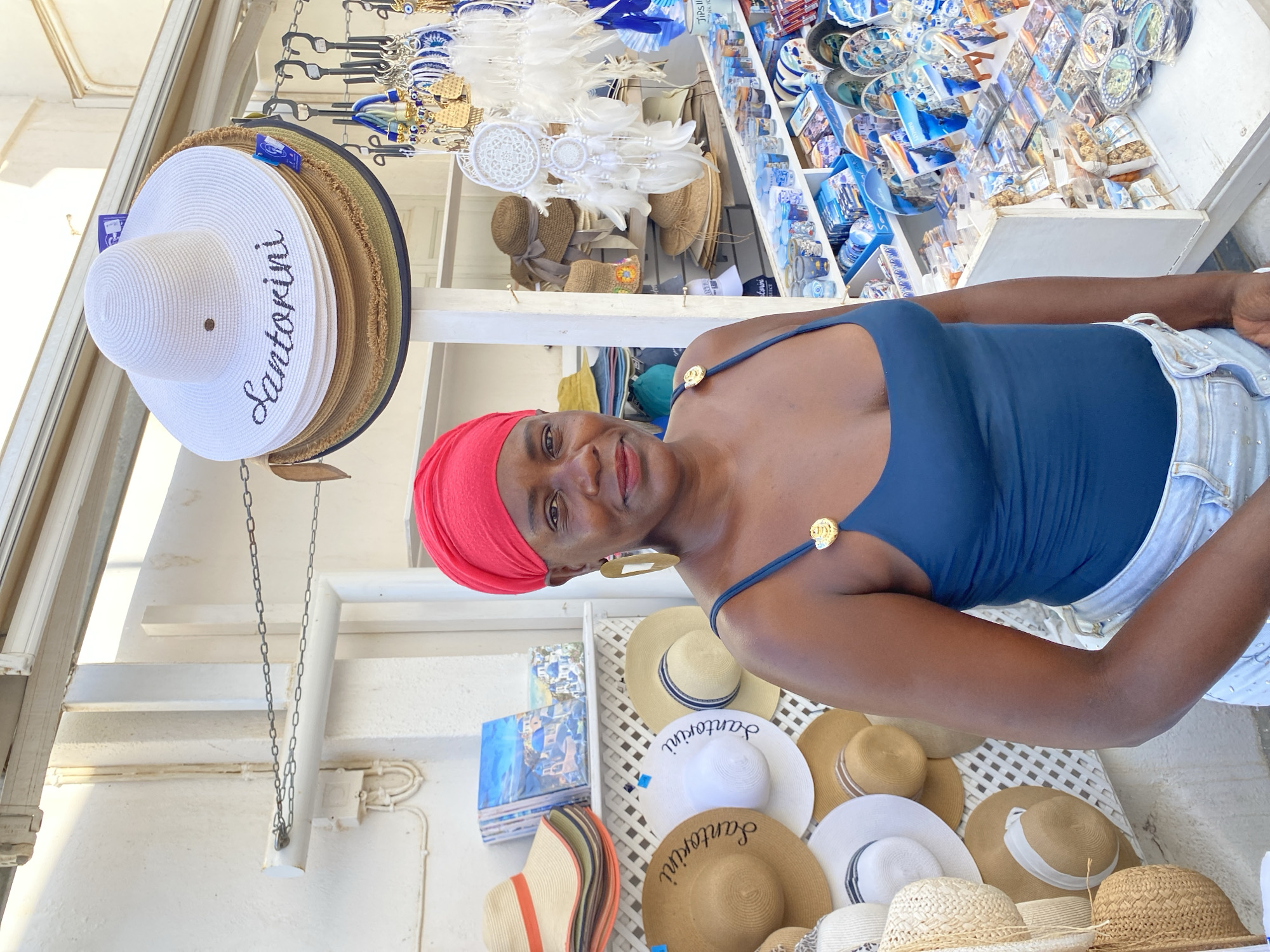 A woman with dark skin, wearing a red headscarf and a dark blue swimsuit, stands in front of a shop stall in the city of Ia, Greece. The stall displays various souvenirs, including hats, dreamcatchers, and small plates featuring images of white buildings with blue domes, characteristic of the Cycladic islands. The woman is looking towards the camera with a slight smile. Behind her, a selection of straw hats, some with "Santorini" written on them, are displayed on a white lattice wall. The shop appears to be outdoors, with a white building and awning visible in the background.
