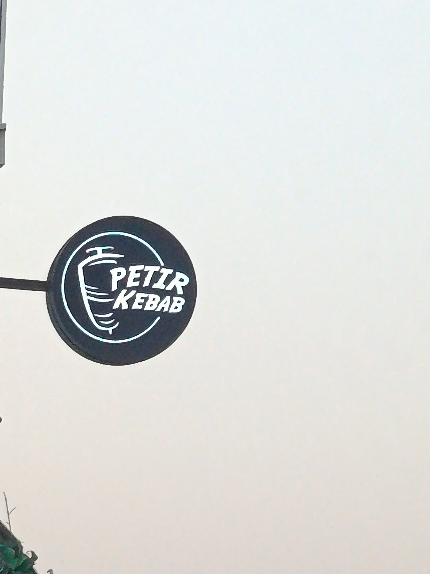 A close-up, outdoor shot shows a black circular sign with white lettering and graphics against a pale, overcast sky. The sign is angled, with a black pole extending from its left side. The graphic on the sign depicts a stylized spit with meat. The text on the sign reads "PETIR KEBAB" in white, block capital letters. The sign appears to be illuminated from within or reflecting light, giving it a bright outline. The bottom left corner of the image shows a dark, out-of-focus area with green foliage, possibly indicating a plant or tree. The overall impression is of a street sign for a food establishment.