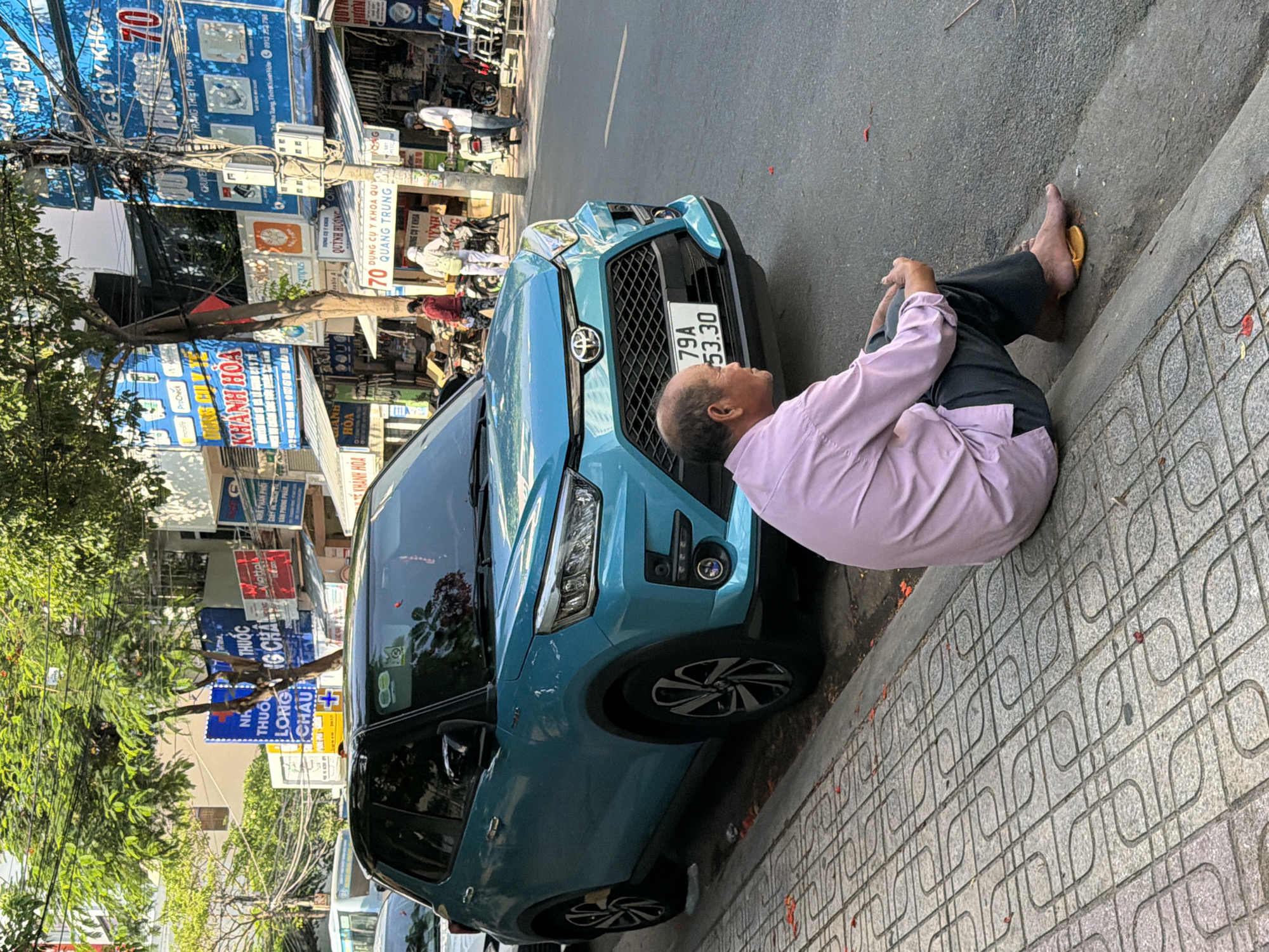 In Nha Trang, Vietnam, an older man in a light pink button-down shirt and dark pants sits on the curb on a sunny day, facing a light blue SUV parked on the street. The man is barefoot with one yellow sandal placed near his feet. The SUV has a license plate displaying "79A 53.30". Behind the car, various shops and signs are visible, including "Y NHÀ THUỐC LONG CHÂU" and other businesses with Vietnamese text. Trees with green foliage hang over the sidewalk, and power lines are strung across the scene. The sidewalk is paved with patterned grey tiles, and the street is asphalt. Several motorcycles are parked along the roadside in the background, and other pedestrians are present further down the street.