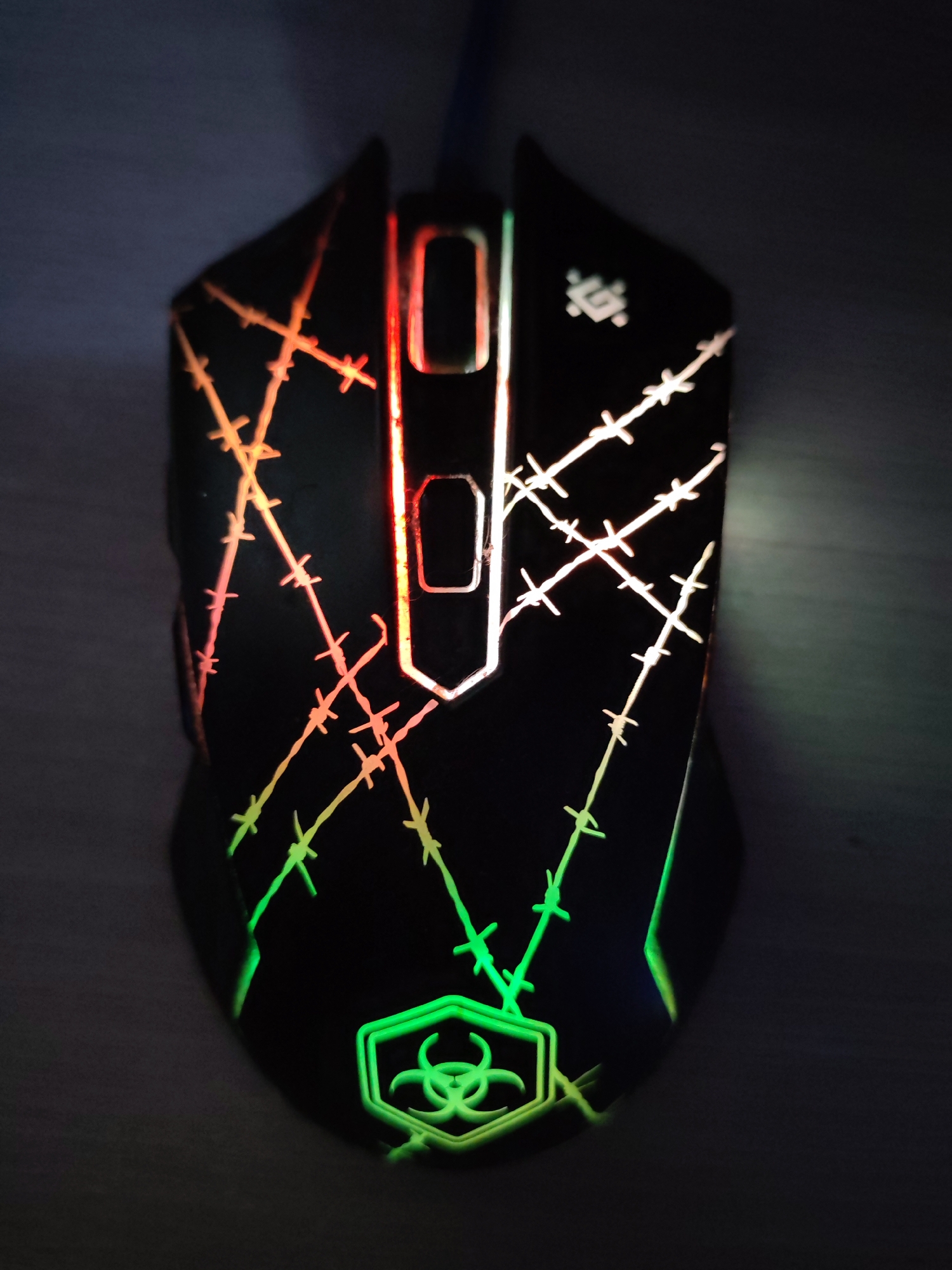 A top-down view depicts a black computer mouse with multi-colored LED lighting. The mouse features a decorative pattern resembling barbed wire in shades of orange, white, and green. Near the bottom, there's a green biohazard symbol. The mouse has two illuminated buttons in the middle, lit in red and white. The image background is a dimly lit surface, likely a desk. The location of Chaikovsky, Russia, cannot be verified from the image.