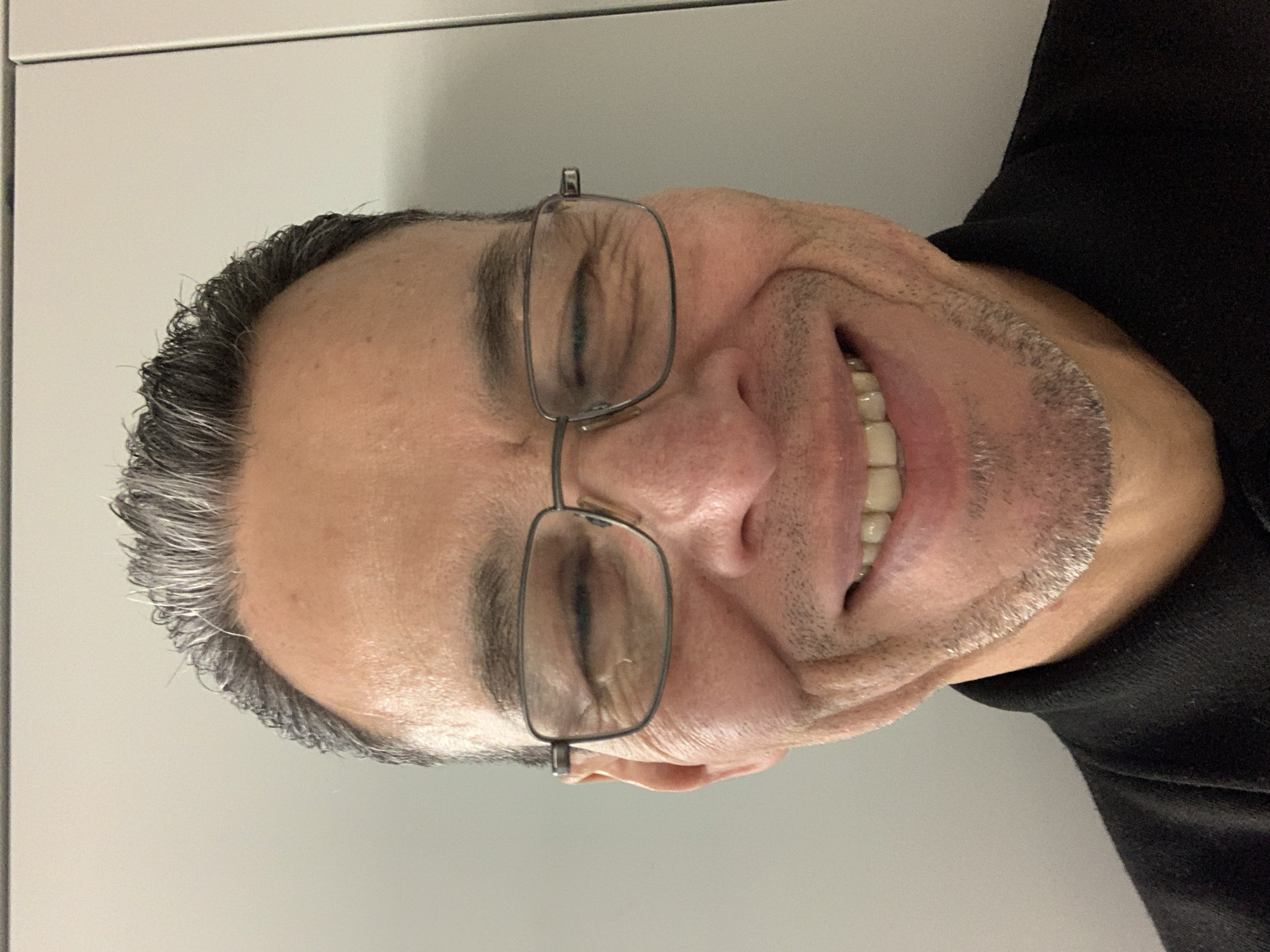 A close-up, slightly out-of-focus photograph depicts a man with graying hair and stubble, wearing glasses. He is smiling with his eyes closed. The background is a plain, light gray wall. The man is wearing a dark-colored garment. There is no indication in the image that the location is London, United Kingdom.