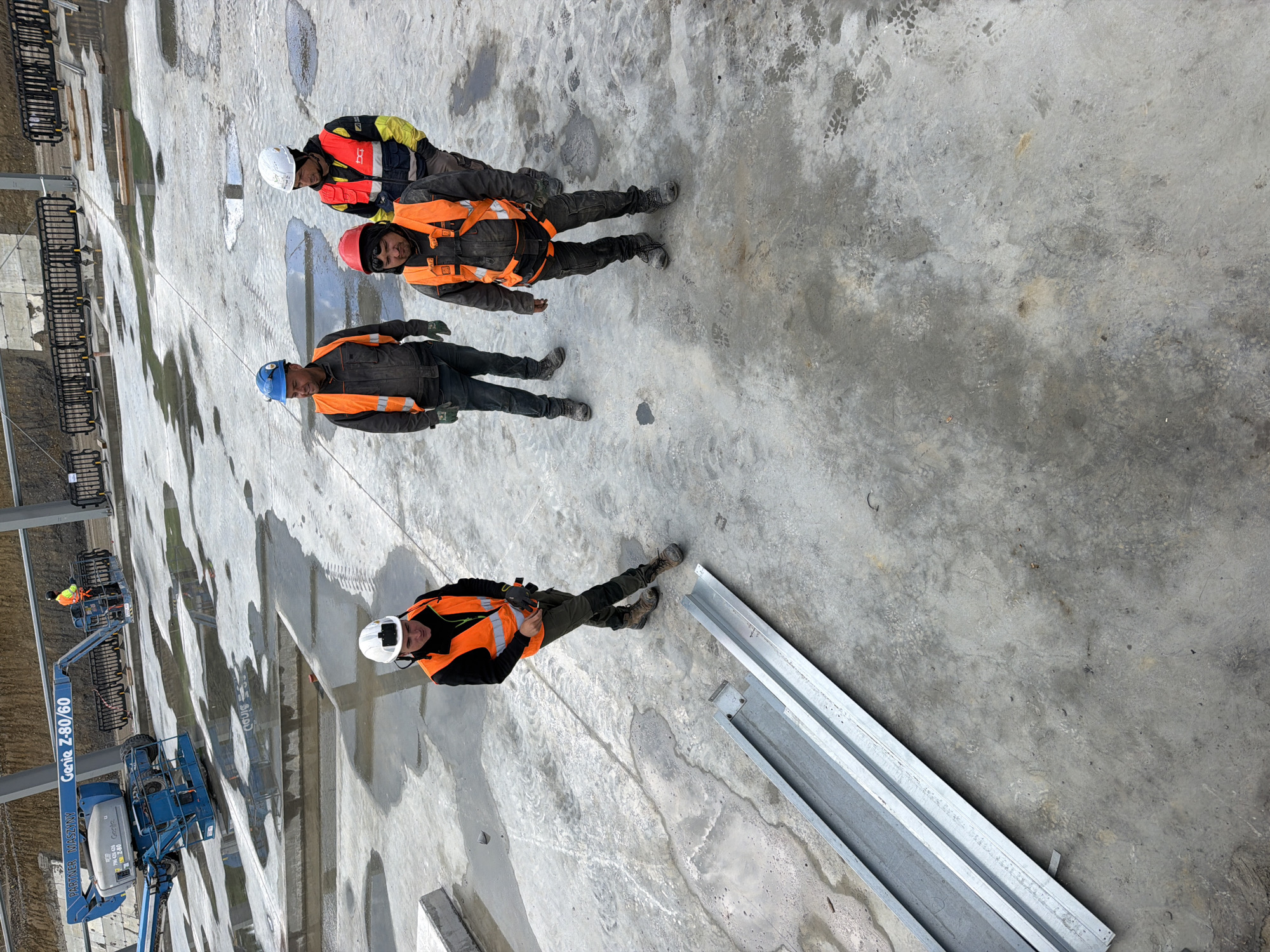In Stróża, Poland, four construction workers are gathered on a wet, grey concrete surface. The workers are wearing hard hats and high-visibility orange vests over their work clothes. The worker on the far left is wearing a white hard hat and an orange vest, looking down towards the ground. The worker next to him, to the right, is wearing a blue hard hat and a dark jacket with an orange vest. He is looking to his left. In the center, a worker with a red hard hat and an orange vest is looking forward. The worker on the far right is wearing a white hard hat and a yellow and black vest over an orange one, also looking forward. In the background, a blue aerial work platform is parked. The ground is covered in puddles of water and tire tracks, indicating recent activity. A long, metallic construction beam lies on the concrete in the foreground.