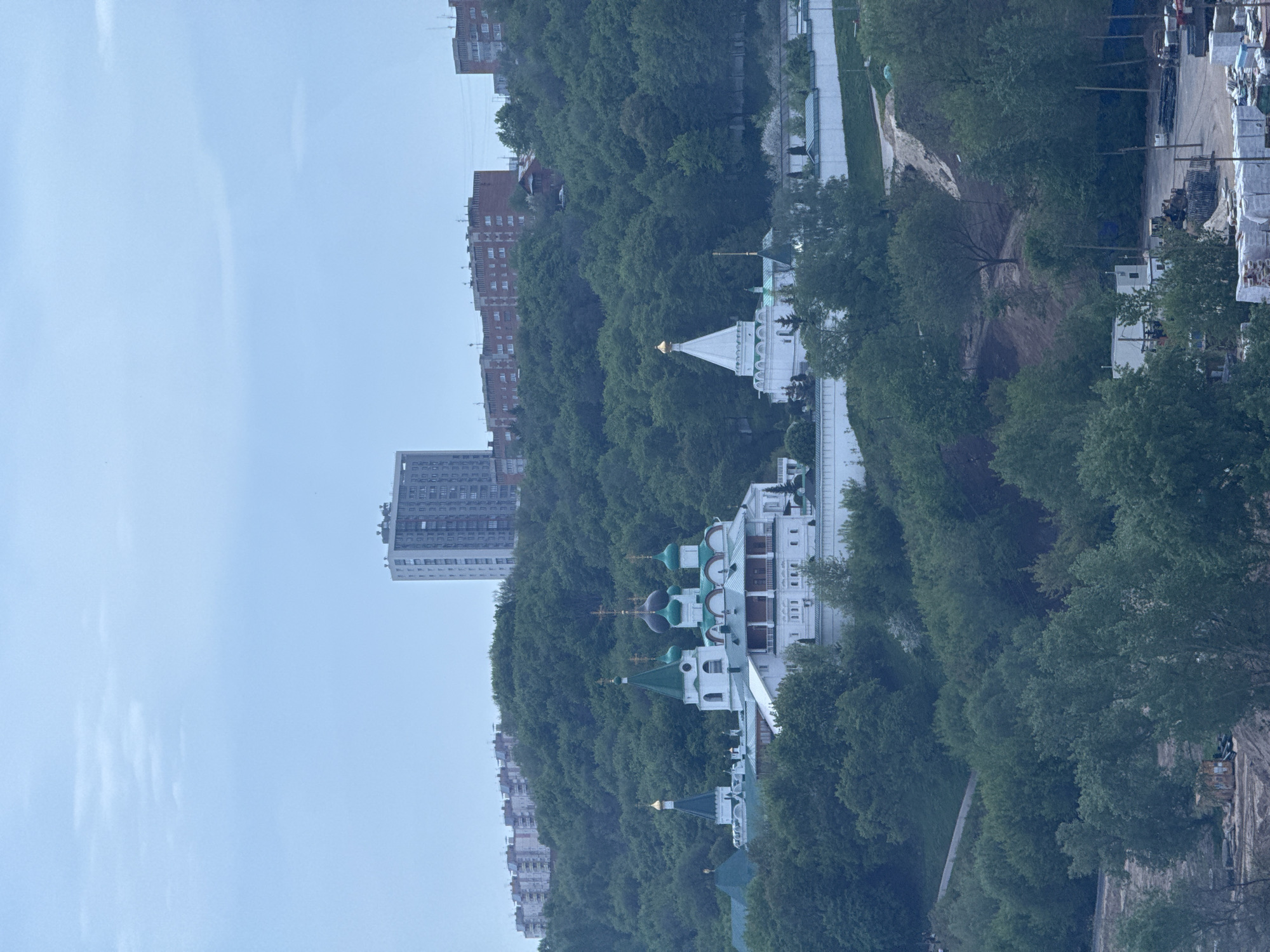 The image shows a vertical view of a Russian Orthodox monastery complex nestled among lush green trees and hills, with modern buildings in the background. This is located in the city of Nizhny Novgorod, Russia. The architectural style of the monastery features white buildings topped with green and gold domes. A tall, modern apartment building is visible to the left, providing a stark contrast to the traditional religious architecture. The sky is overcast. The general environment suggests an urban area blending with natural elements.