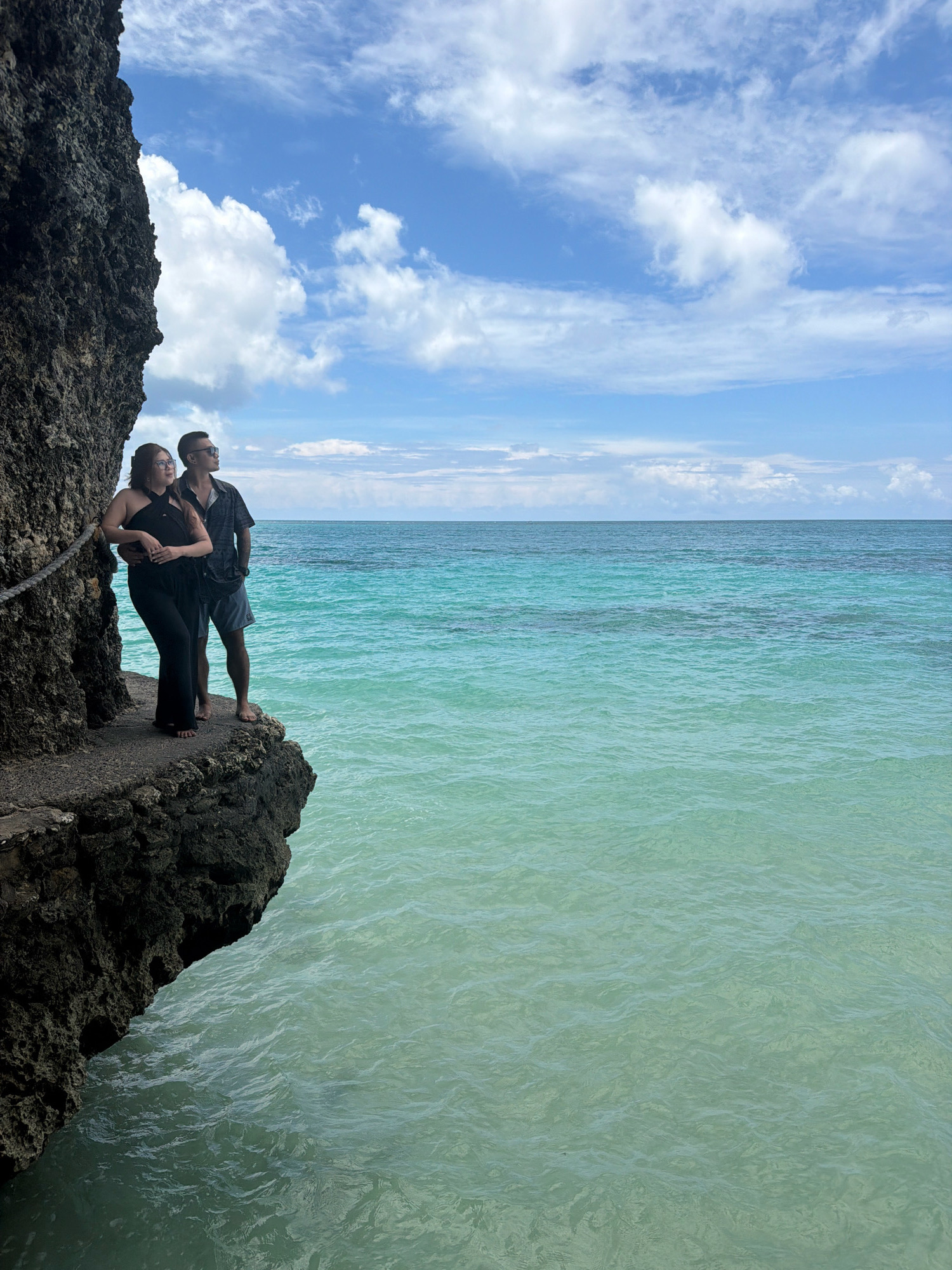 Two people stand on a rocky outcrop overlooking a turquoise ocean under a partly cloudy sky. The male subject, wearing sunglasses, a dark patterned short-sleeved shirt, and light gray shorts, has his arm around the female subject. The female subject, wearing a black jumpsuit and glasses, leans into him. A rope railing is visible on the left side of the outcrop, bordering a sheer rock face. The location is identified as Malay, Philippines.