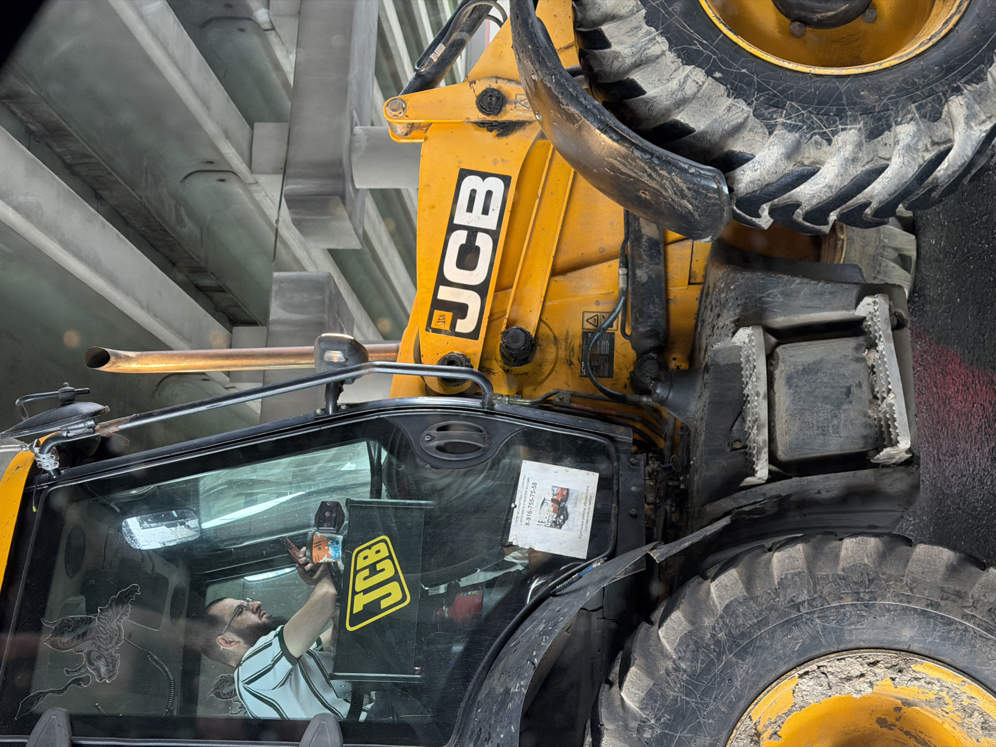 The image depicts a yellow JCB backhoe loader oriented vertically. Inside the cab, a man wearing a white shirt with black stripes and glasses is holding a small orange and blue object. A flyer is attached to the front windshield, partially obscuring the view. The JCB logo is visible on the machine's arm and the cab door. The loader is surrounded by a concrete structure with visible beams. The location is the city of Unknown, Unknown.
