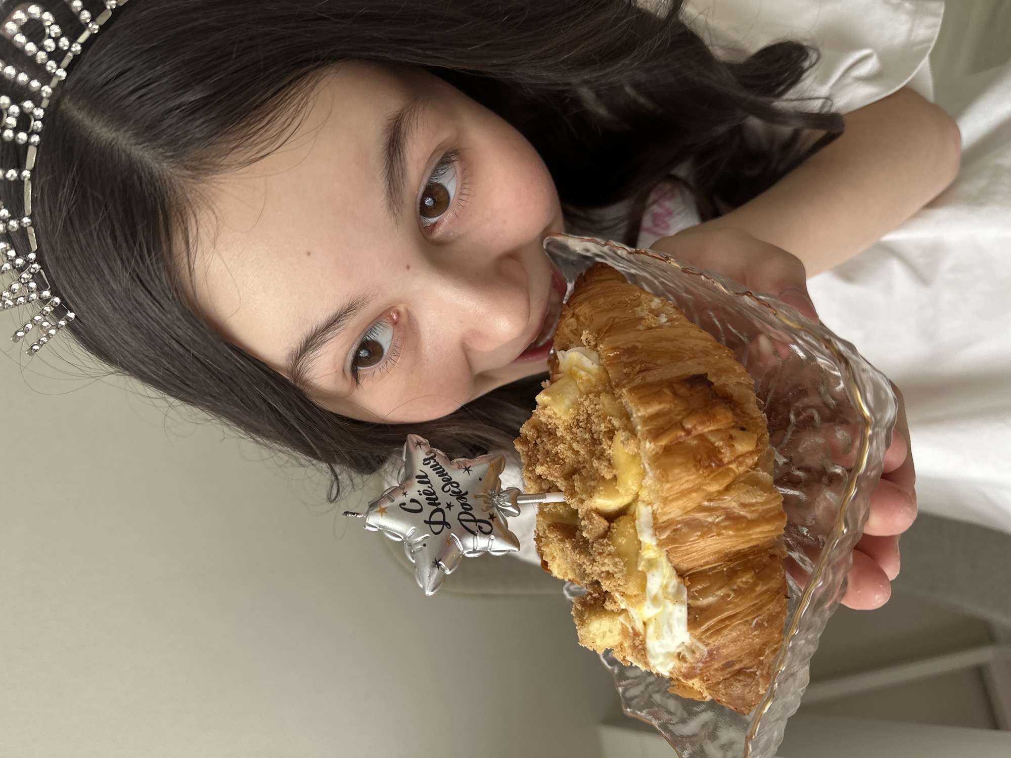 A young person with dark hair, wearing a tiara, is holding a croissant with filling and a star-shaped candle in a decorative wrapper. The candle has writing in a language that appears to be Russian. The person is looking at the camera and about to eat the croissant. The background appears to be indoors with light-colored walls and bedding. There is no clear evidence of the location being Votkinsk, Russia in the image.