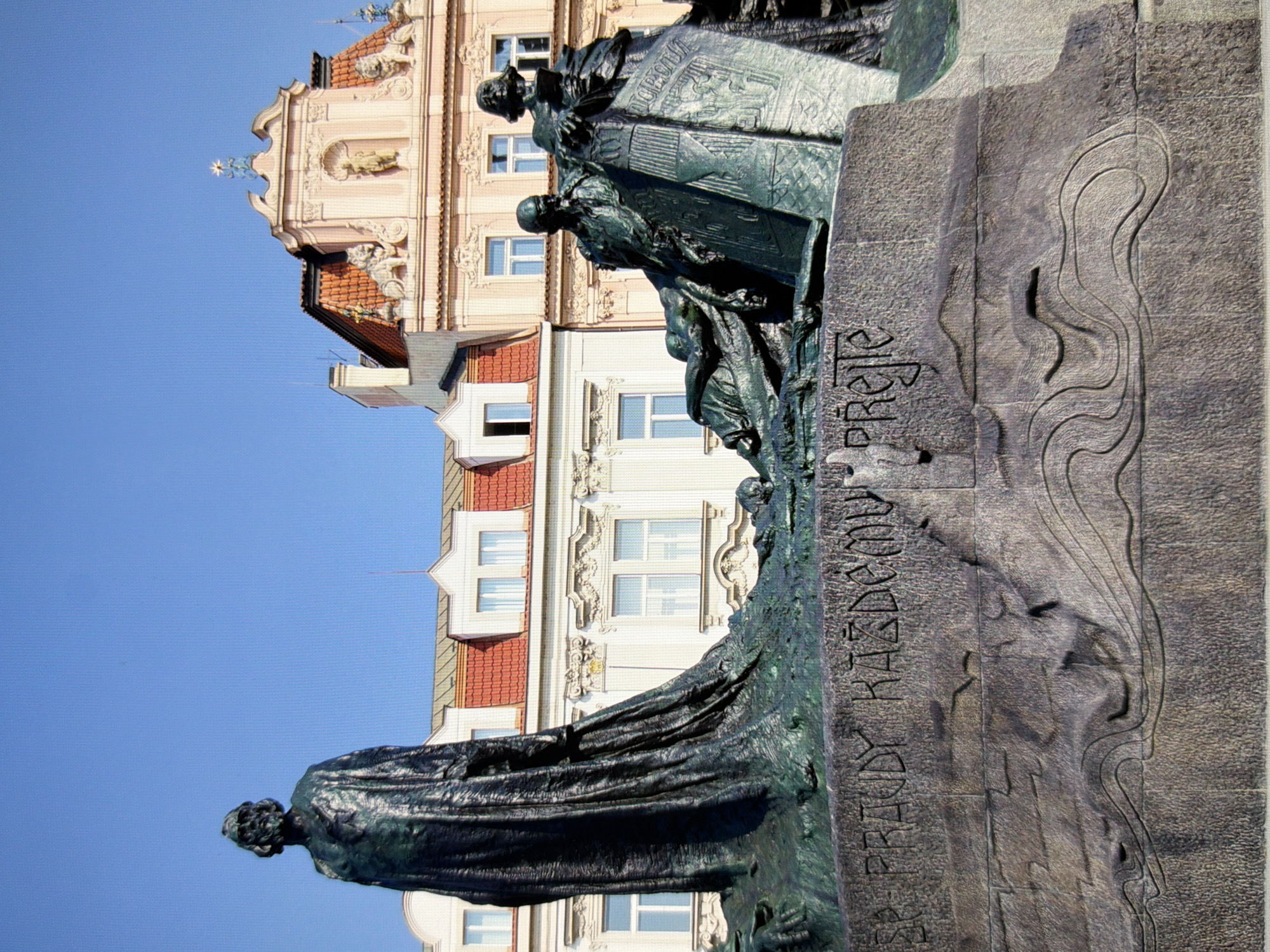 The image shows the Jan Hus Memorial in Prague, Czech Republic. It is a large bronze monument featuring several figures and inscriptions, with a backdrop of historic buildings and a clear blue sky. The monument depicts Jan Hus, a religious reformer, with other figures and allegorical elements. The inscription on the base of the monument is partially visible. The architectural style of the surrounding buildings suggests a European city, consistent with Prague.
