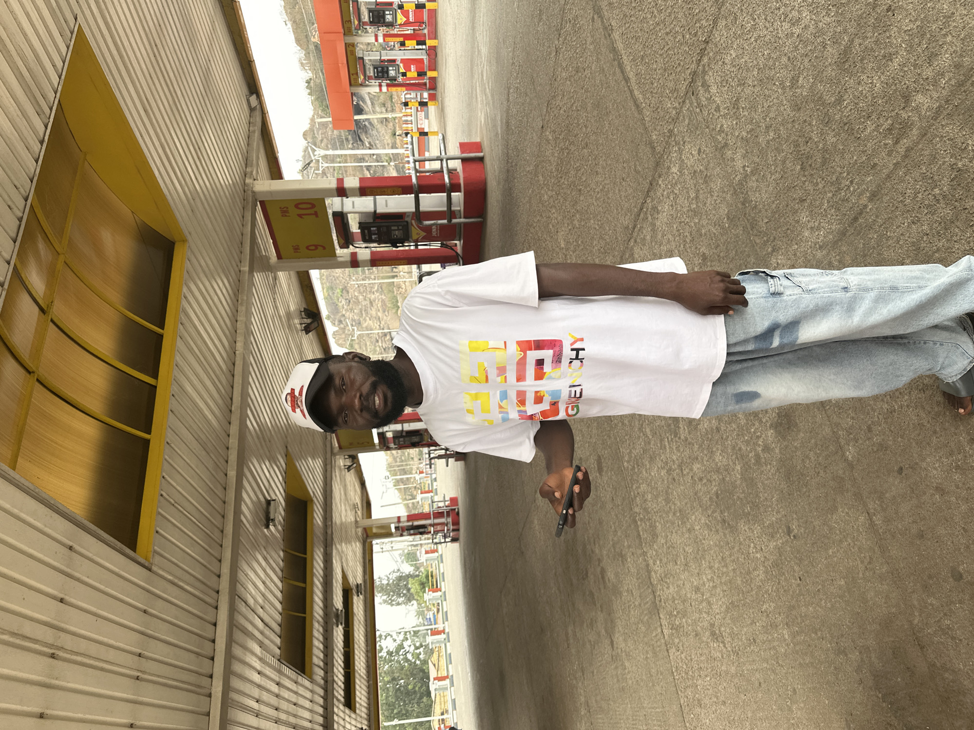 The image captures a man standing at a gas station, appearing to be in Jos, Nigeria, during daylight hours. The man, with dark skin and a beard, is positioned slightly off-center and looks directly at the camera with a faint smile. He is dressed in a white t-shirt featuring colorful, block-style lettering that spells "GIVENCHY" across the chest. He also wears light blue jeans, dark sandals, and a white baseball cap with red accents. In his right hand, he holds a dark smartphone.
The scene is an outdoor petrol station with multiple red and black gas pumps visible behind him, some displaying the "JENNY" brand logo. A white sign near the pumps clearly shows "PMS 9" and "PMS 10," likely referring to pump numbers or fuel specifications. To the man's left is a building with light-colored corrugated metal siding and distinct yellow-framed windows. The ground is a textured light gray pavement. In the distance, beyond the gas station infrastructure, hills or mountains can be seen under a bright sky, indicating an elevated or rugged landscape. The overall atmosphere is relaxed and captures an ordinary moment at a fueling station.