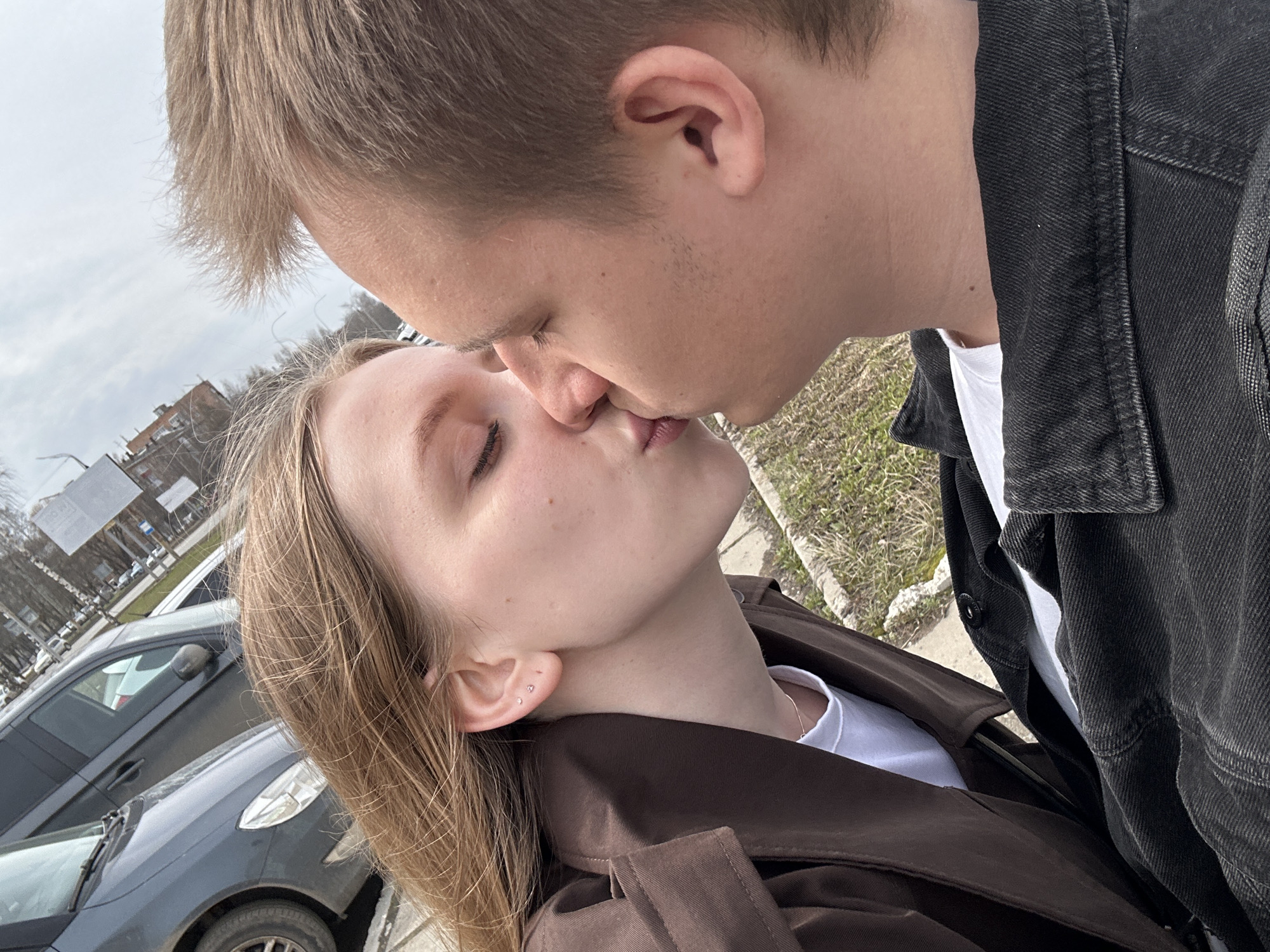 Two people, a man and a woman, are kissing outdoors. The woman has blonde hair and is wearing a brown coat over a white shirt. She has her eyes closed. The man is also blonde and wearing a dark denim jacket over a white shirt. In the background, there are cars and buildings, indicating an urban environment. The location is identified as Izhevsk, Russia.