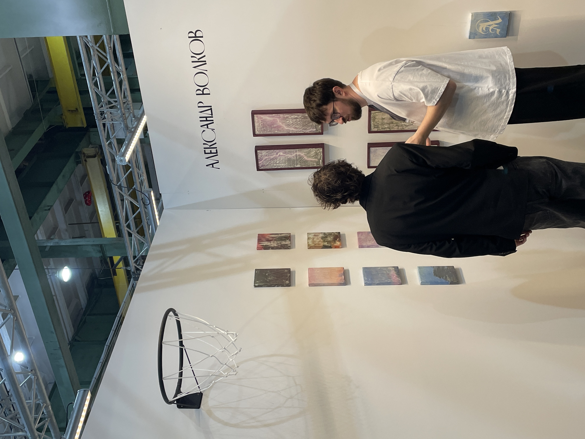 Two people are examining artwork on a white wall. The taller person, wearing a white t-shirt and black pants, is leaning in to observe a series of framed, rectangular pieces. The second person, wearing a black jacket and gray pants, is also viewing art, specifically a row of square paintings. The name "АЛЕКСАНДР ВОЛКОВ" is printed in black vertically on the upper left corner of the wall. Above the taller individual, another small painting can be seen. Below the row of square paintings is a black basketball hoop. The background includes industrial elements: visible metal beams and structures with yellow components, suggesting an interior space reminiscent of a renovated factory or warehouse. The setting appears to be in an art gallery or exhibition. I am unable to confirm the location is Saint Petersburg, Russia based on the visual data.