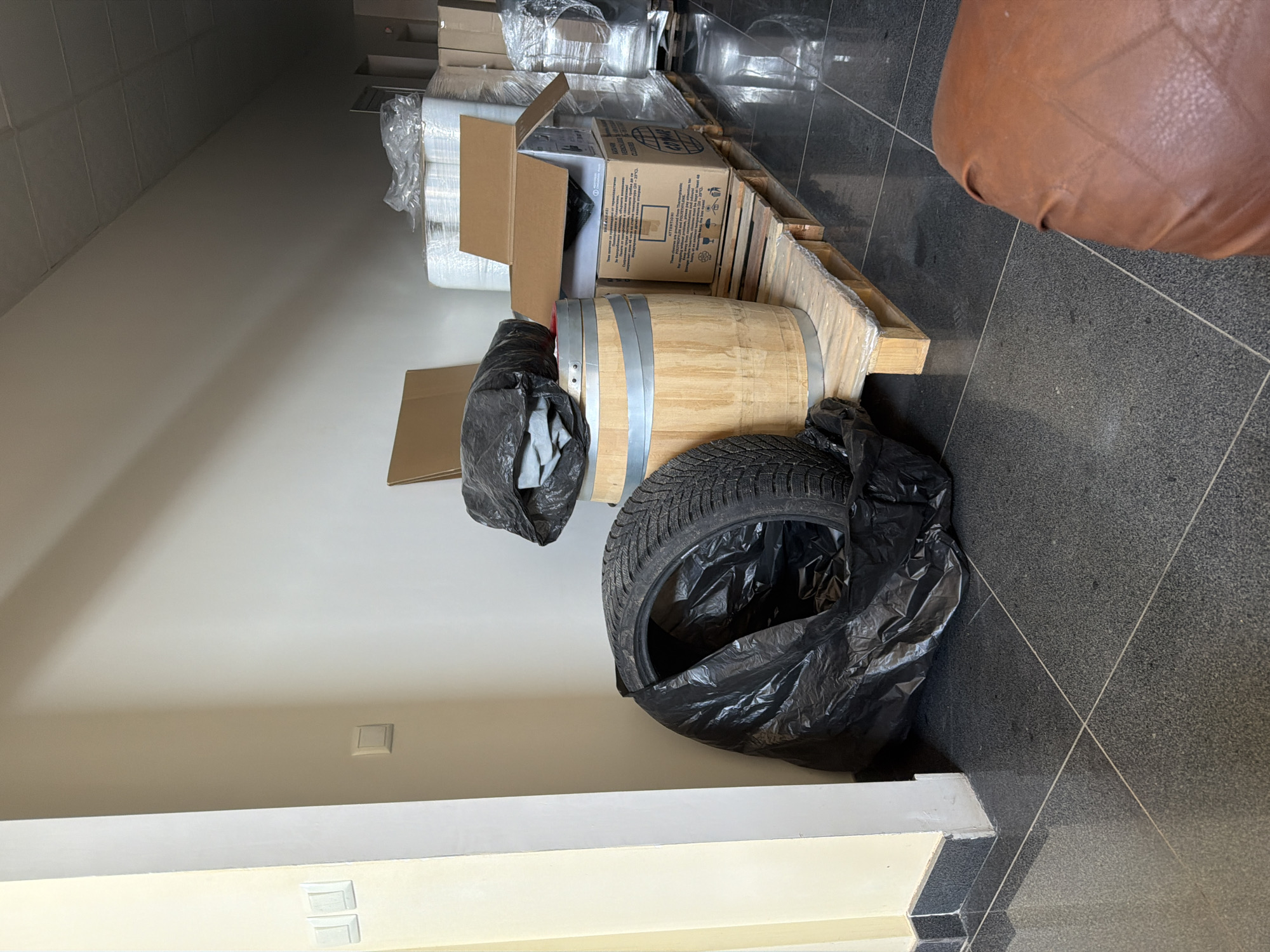 The media file depicts a cluttered indoor scene, likely a storage area or workspace, with a focus on various objects. In the foreground, a large black plastic bag containing a tire and other items sits on the dark, speckled tile floor. Beside it, a wooden barrel with metal bands is partially visible. Behind the barrel, a cardboard box is open, revealing more packing materials inside. Further back, stacks of items wrapped in clear plastic and more cardboard boxes are arranged on wooden pallets. The lighting suggests an indoor environment, possibly with artificial light, as there are no obvious indications of natural light or weather conditions. The overall impression is one of disarray, with items piled and stored without apparent organization. No people are visible in the image. The location context provided is Dolno Ablanovo, Bulgaria.