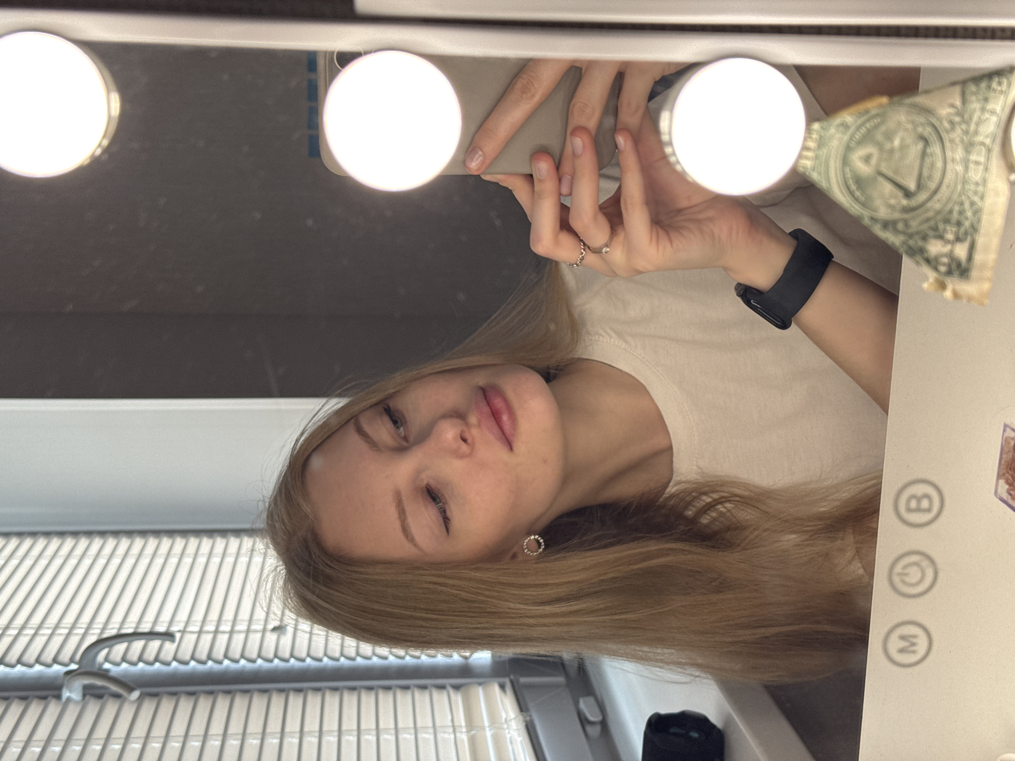 A selfie captured in a bathroom in Perm, Russia. A blonde woman with an earring in her left ear is reflected in a lighted mirror while holding a smartphone. Her right wrist displays a black smartwatch. A US one-dollar bill is partially attached to the top right corner of the mirror frame. A sink with a faucet is visible in the lower left. The mirror frame has buttons marked "M" and "B." The lighting in the scene is bright due to the vanity lights around the mirror.