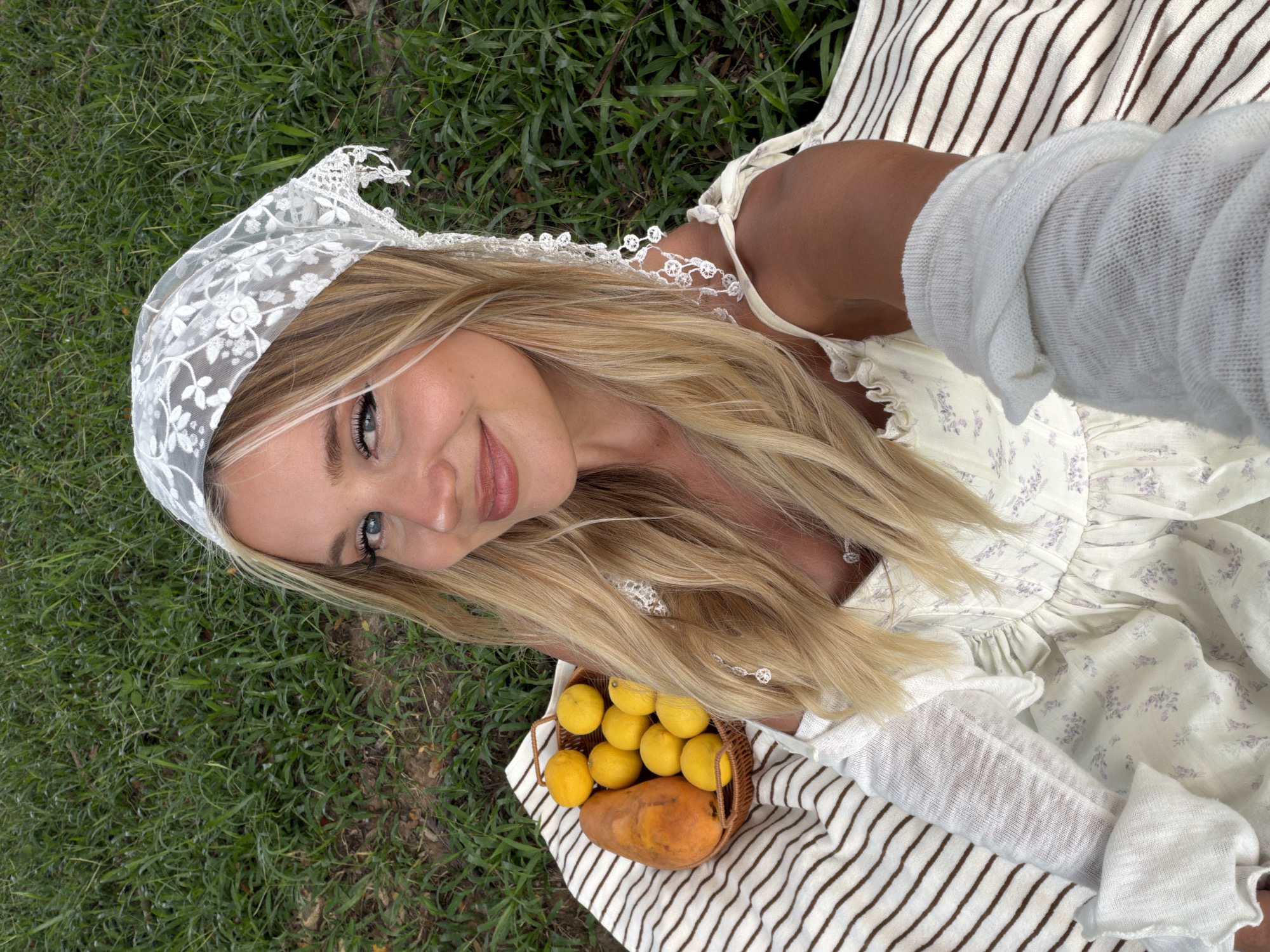 A young blonde woman with blue eyes and a white lace headscarf is taking a selfie while sitting on a striped blanket in a grassy area. She is wearing a white dress with a floral pattern and a white knit sleeve on her right arm. To her left, a woven basket contains several yellow lemons and a papaya. The background is blurred green grass. The scene appears to be outdoors, possibly in a park or garden.