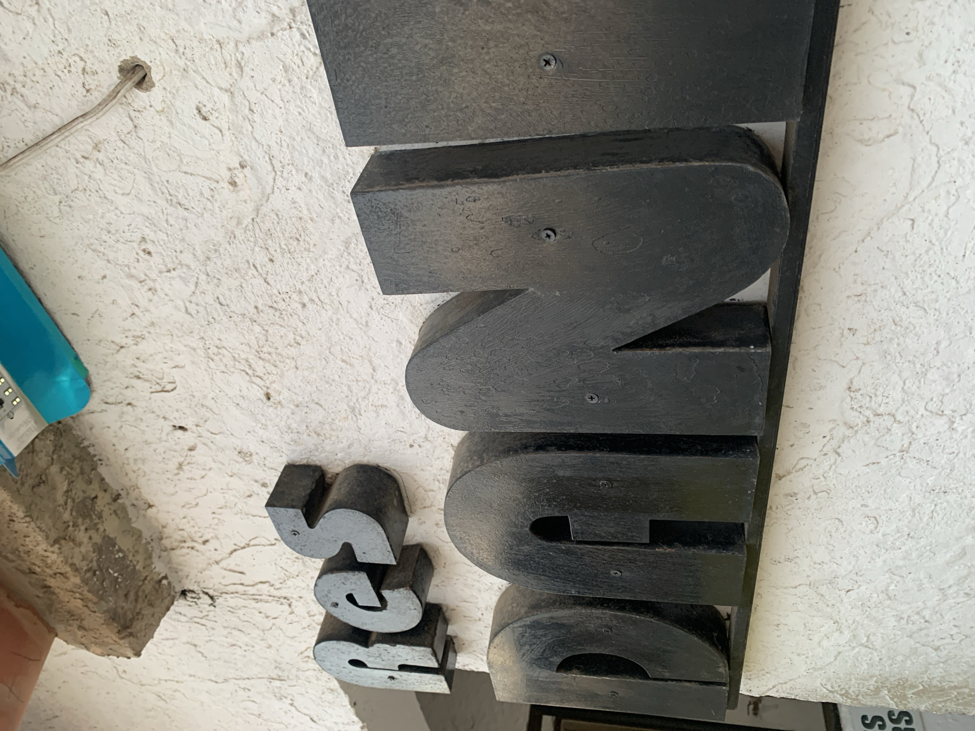 The image displays large, black, three-dimensional letters mounted on a textured white wall. The letters spell out "RES" in the upper portion of the image and "DANI" in the lower portion, suggesting the name "Res Dani". The letters are made of a dark material, possibly wood or metal, and show signs of wear, with visible screws securing them to the wall. Above the "RES" lettering, a rough, concrete-like overhang is visible, with a bright blue object partially in frame at the top right. A wire extends from a hole in the wall to the right of the "RES" sign. The overall scene appears to be an exterior view of a building.
